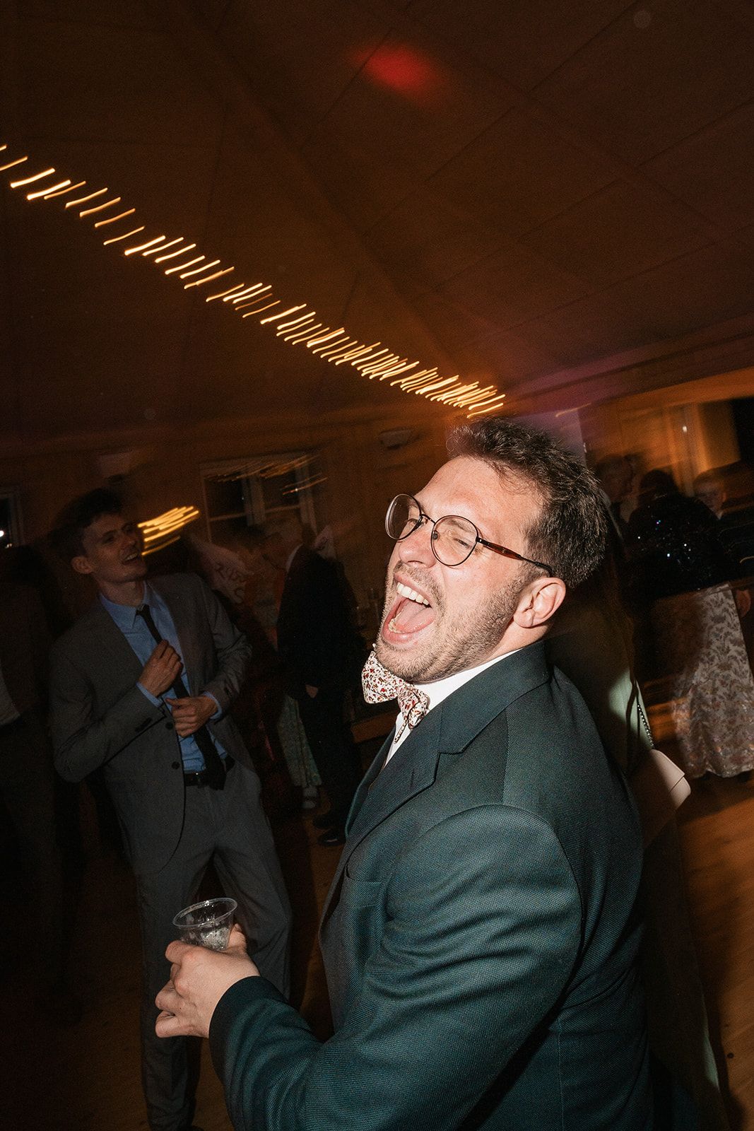 Person in glasses and suit smiles and dances in a dimly lit room at a social gathering.
