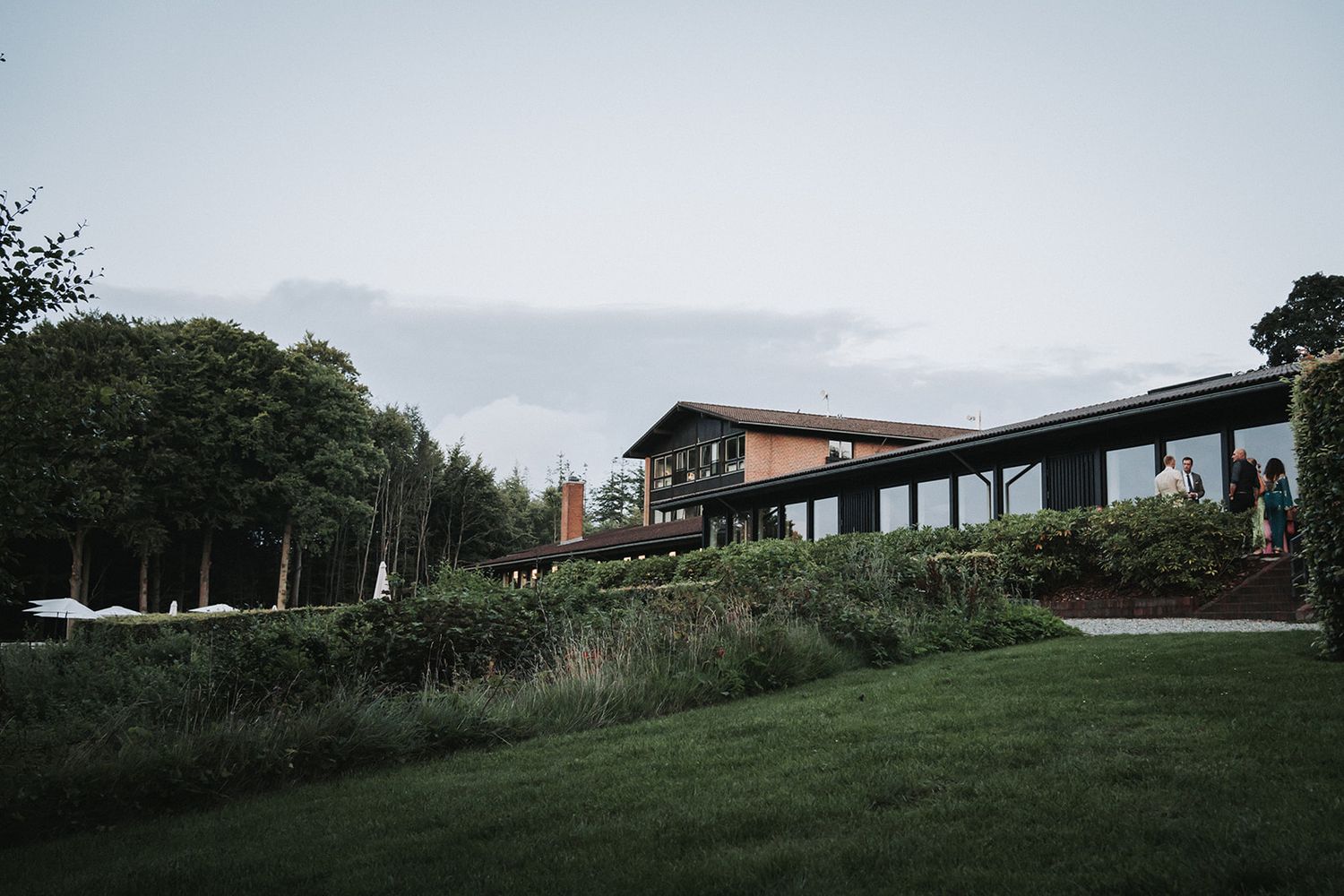 Modern hillside lodge with glass walls and sloped roof situated on a grassy landscape at dusk.