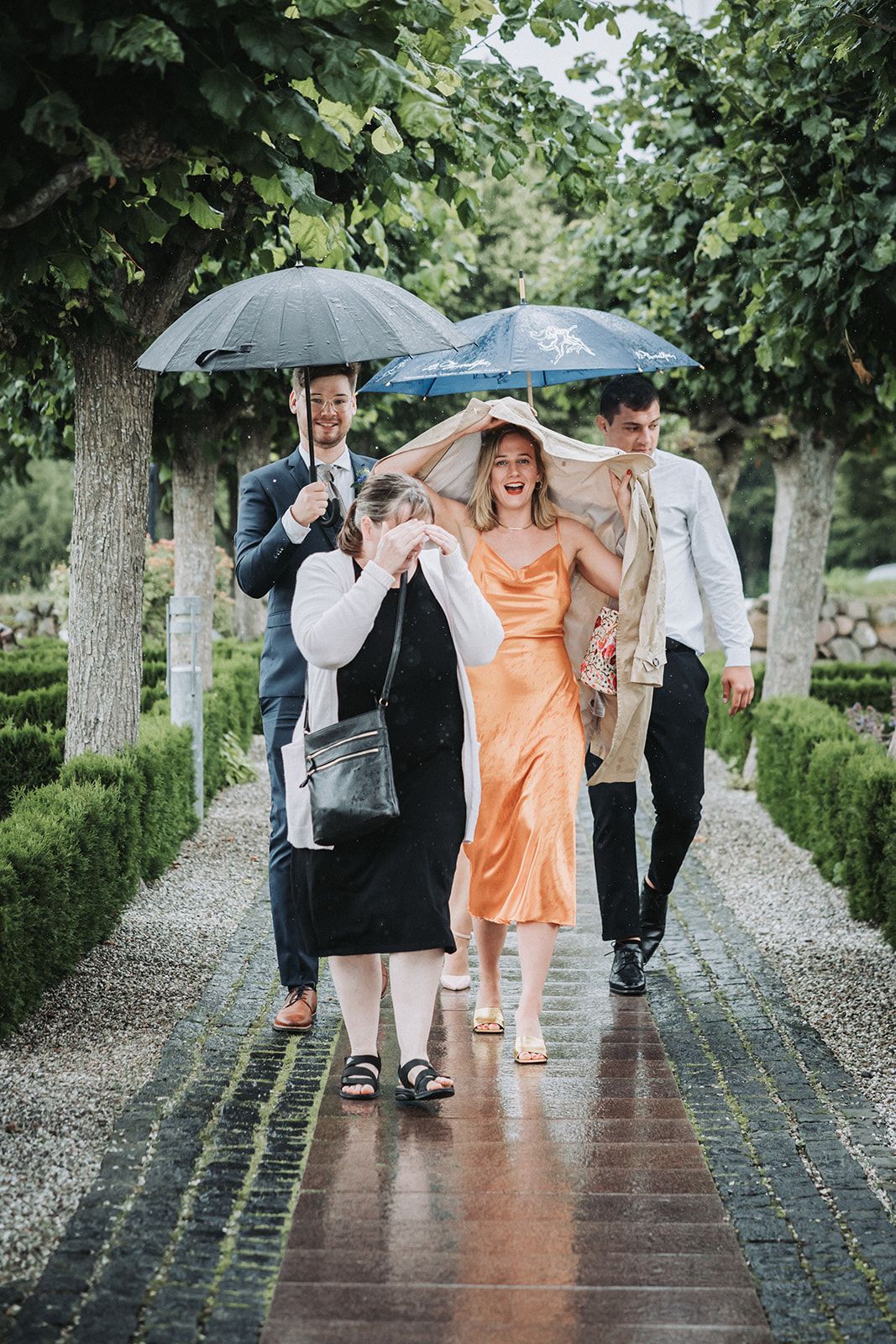 Group walking together with umbrellas on a rainy garden path, woman in peach silky dress laughing in the middle.