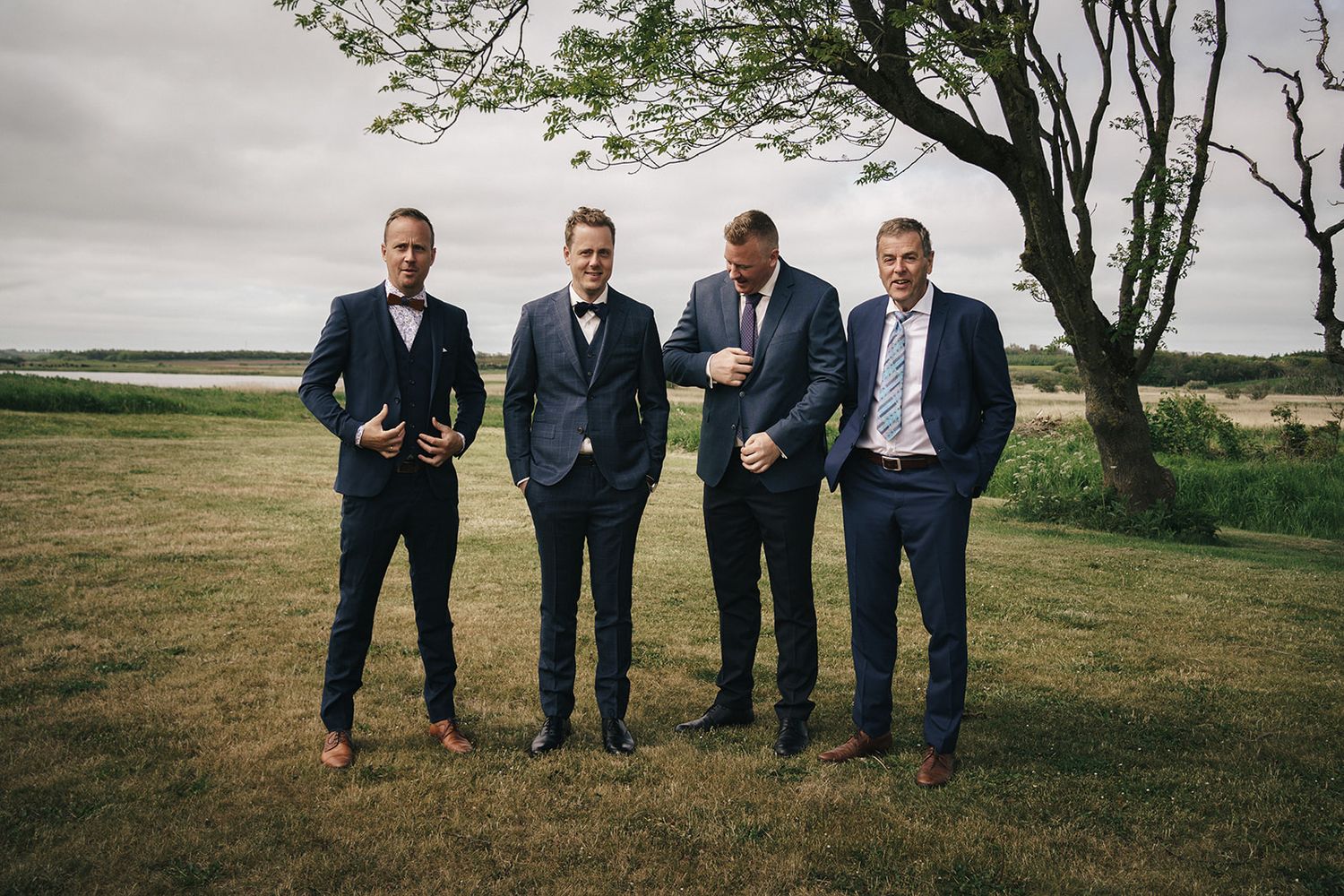 Four men in formal suits stand together on a grassy hilltop overlooking a scenic coastal landscape.