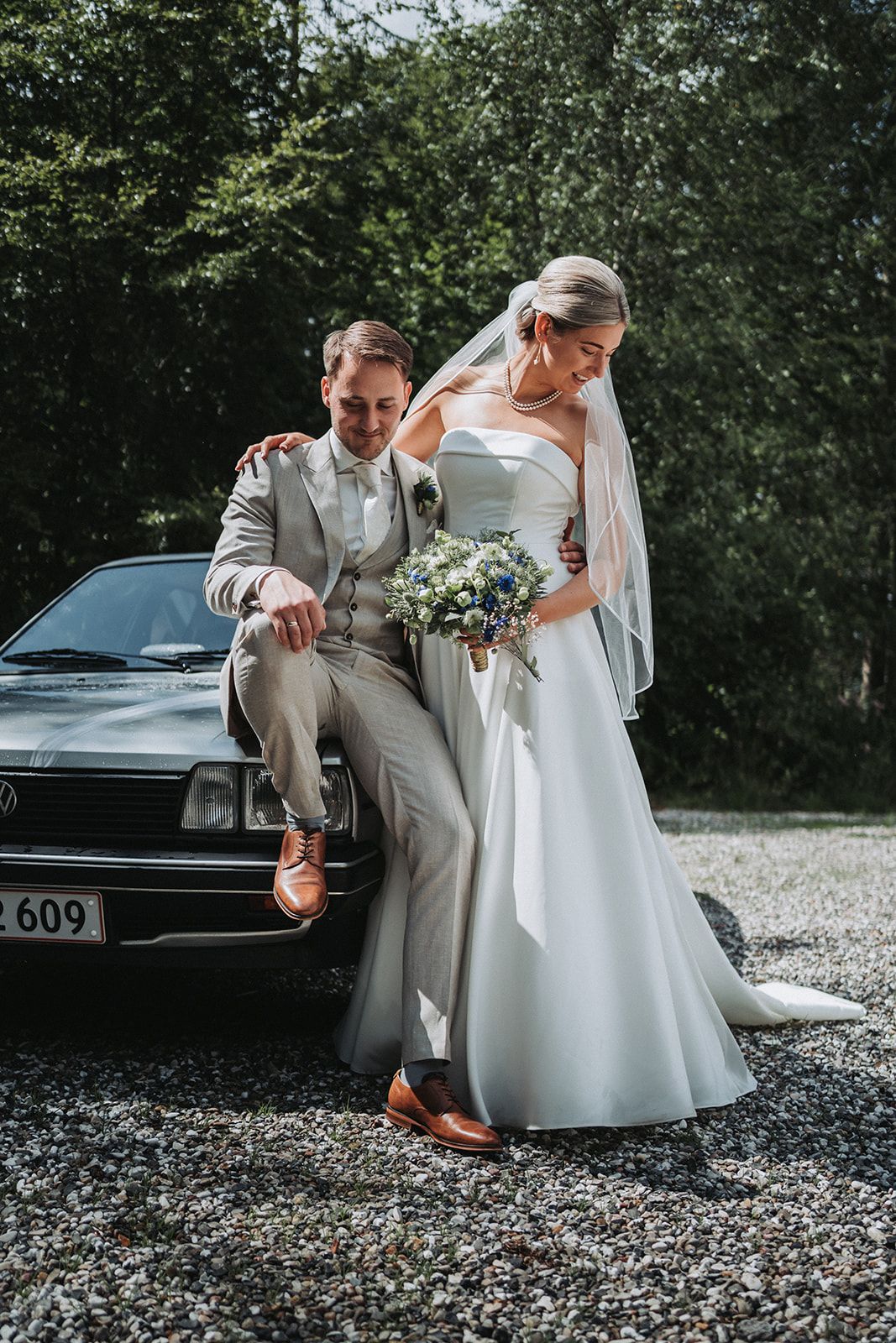 Newlyweds pose together by vintage car with bride holding white floral bouquet and wearing elegant dress.