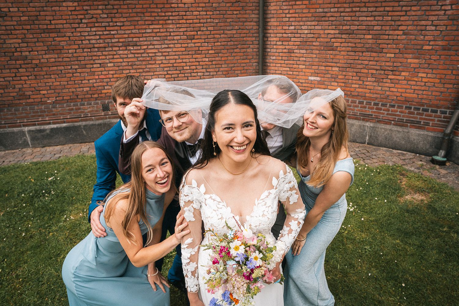 A group of women wearing casual attire and dresses laugh together on a sunny day by a brick wall.