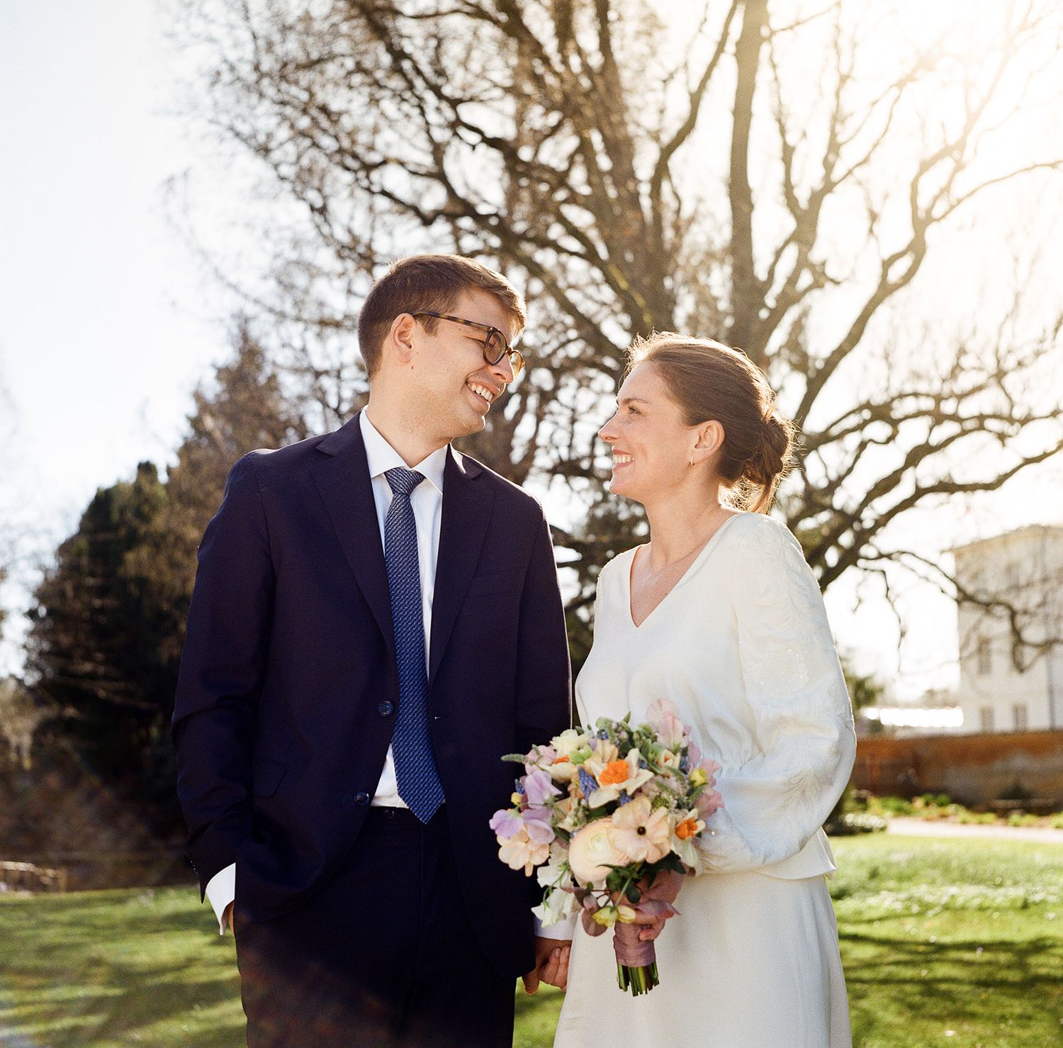 A couple in wedding attire smile at each other outdoors on a sunny day with bare trees in the background.