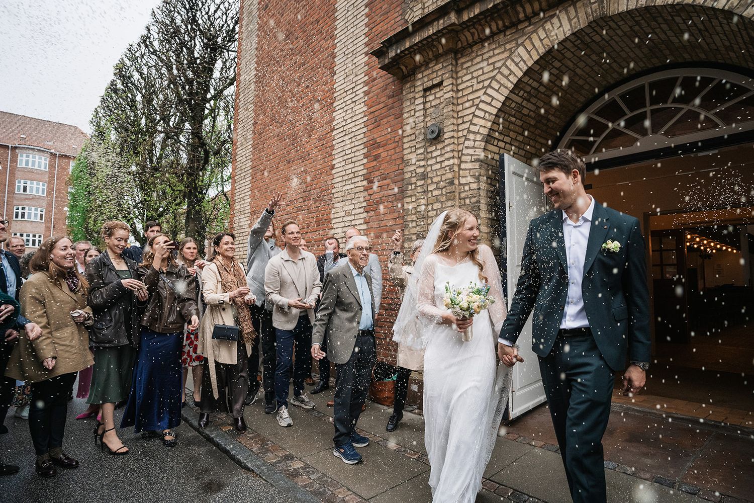Wedding guests shower a newlywed couple with confetti as they exit a brick church building on their special day.