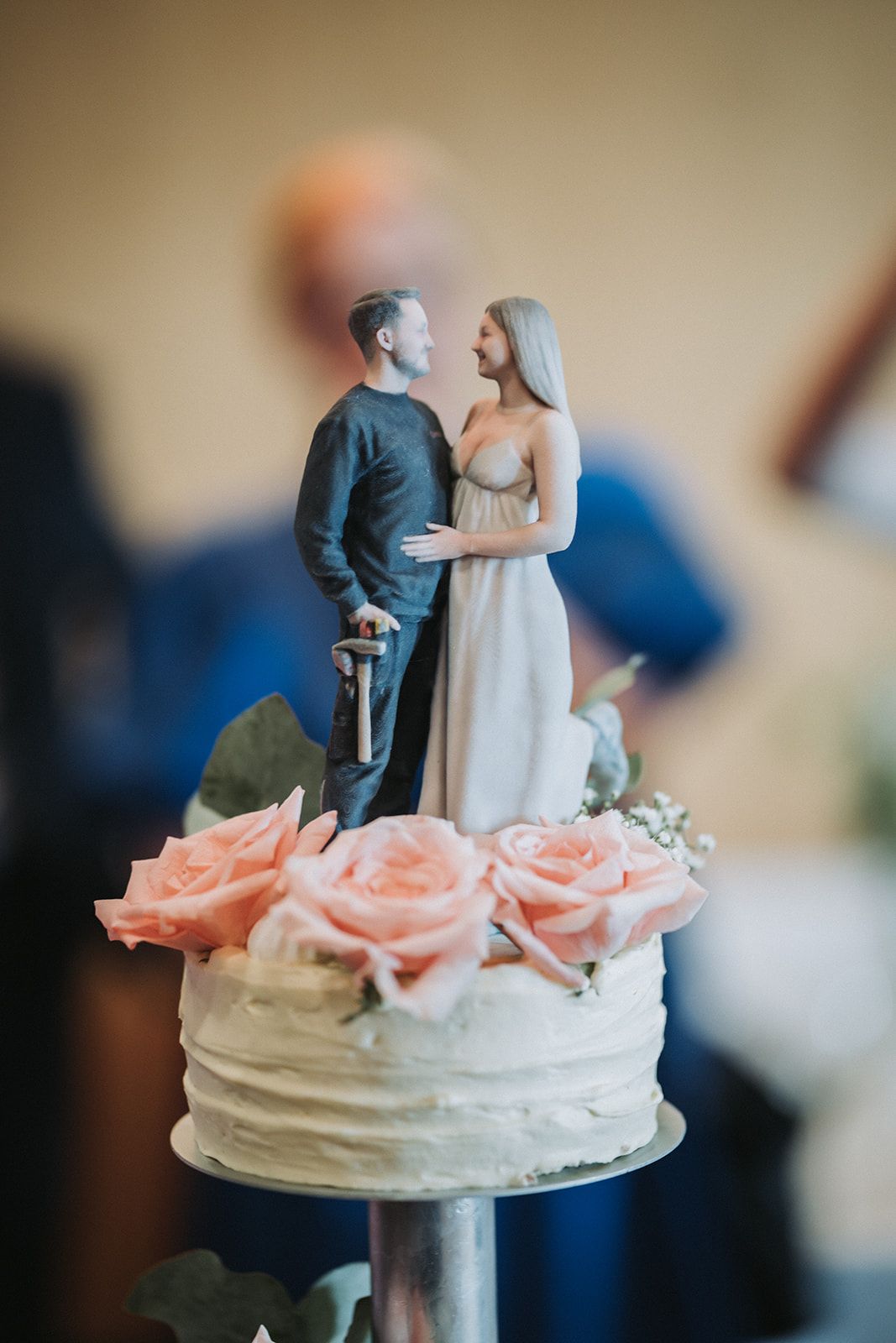 A wedding cake topped with fresh pink roses and a traditional bride and groom figurine topper.