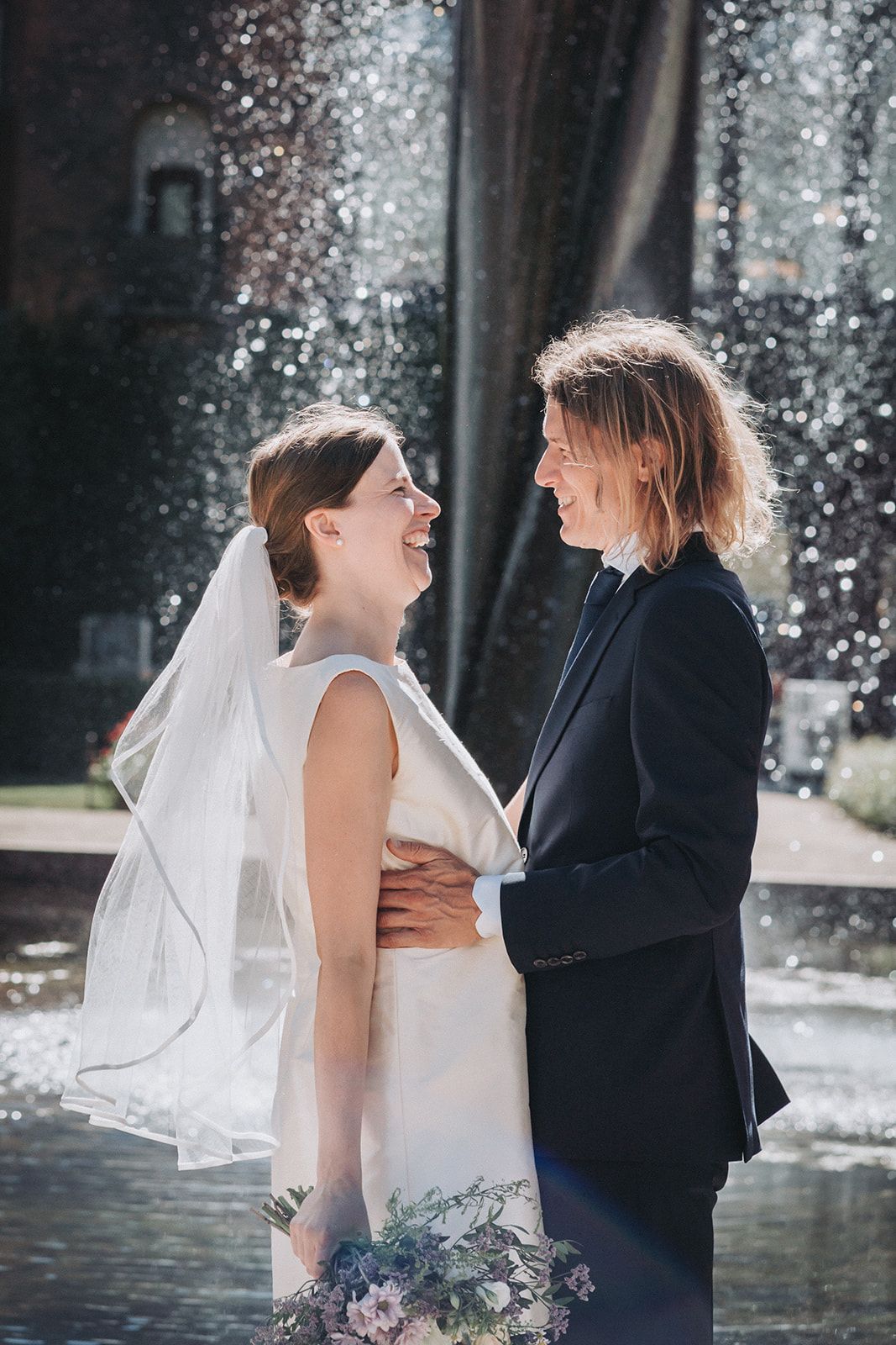 A joyful wedding couple laughs together in front of a fountain, with the bride in white and holding a purple bouquet.