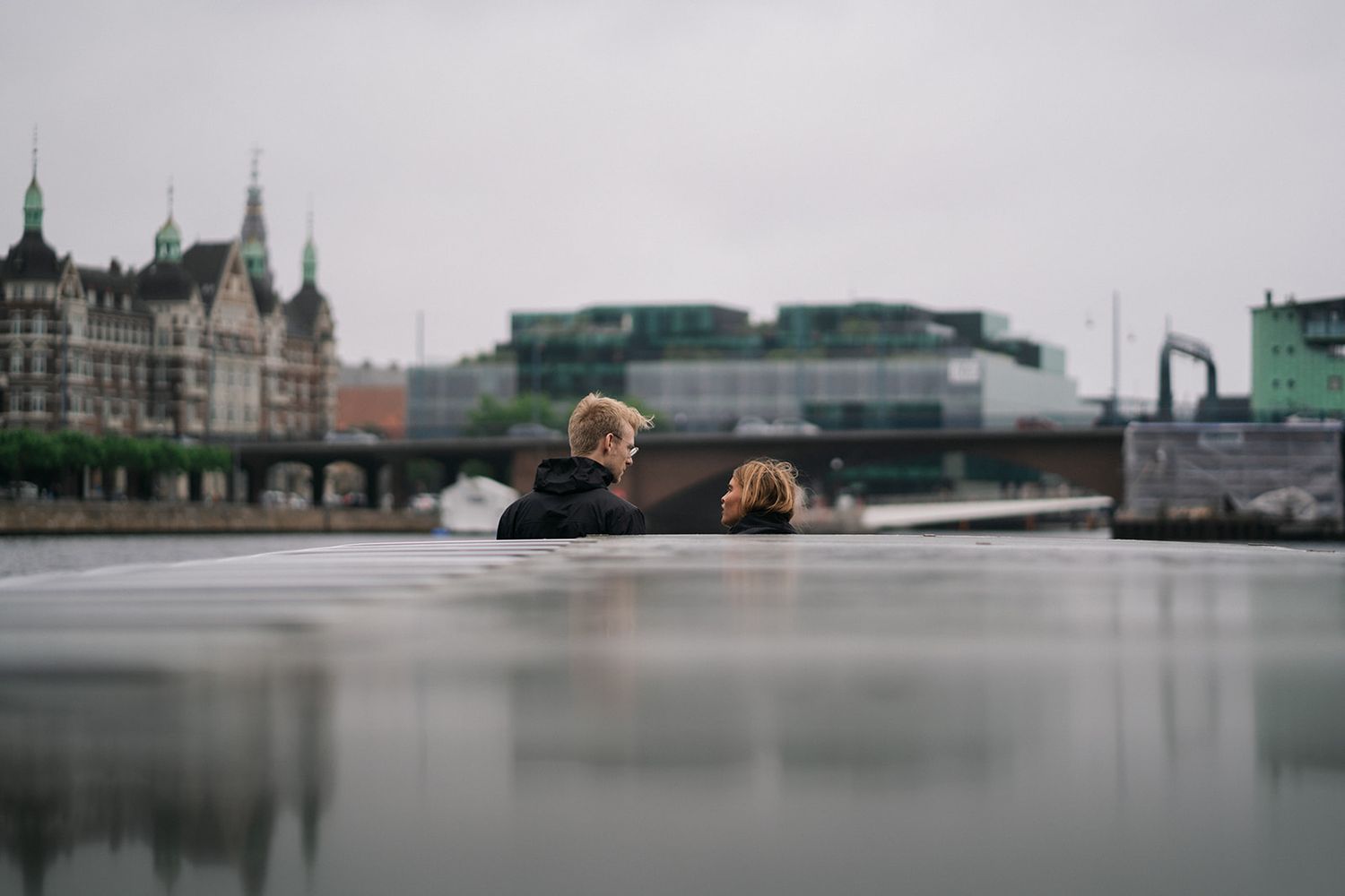 Two people sit by the water's edge overlooking a European cityscape with historic buildings and domes in the background.