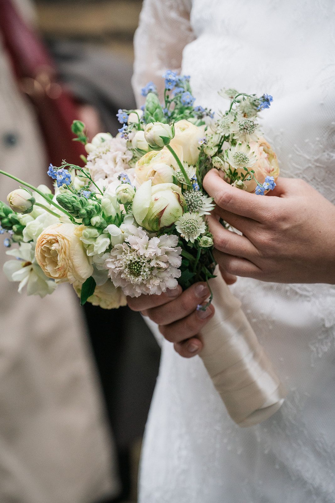 A delicate bridal bouquet with cream roses, white dahlias, and small blue flowers held against a white wedding dress.
