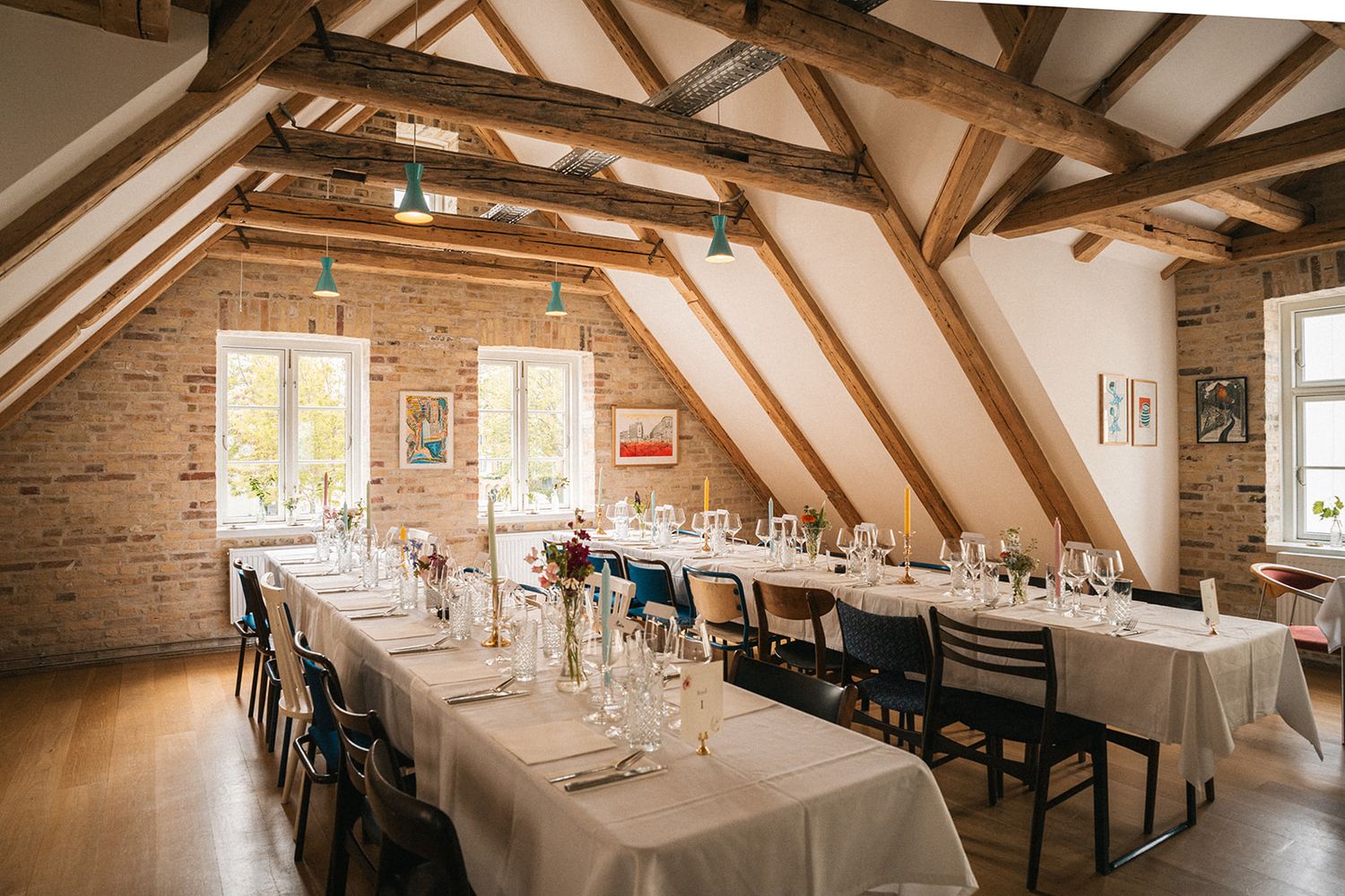 Rustic attic dining room with exposed wooden beams, brick walls and long tables set for an elegant dinner service.