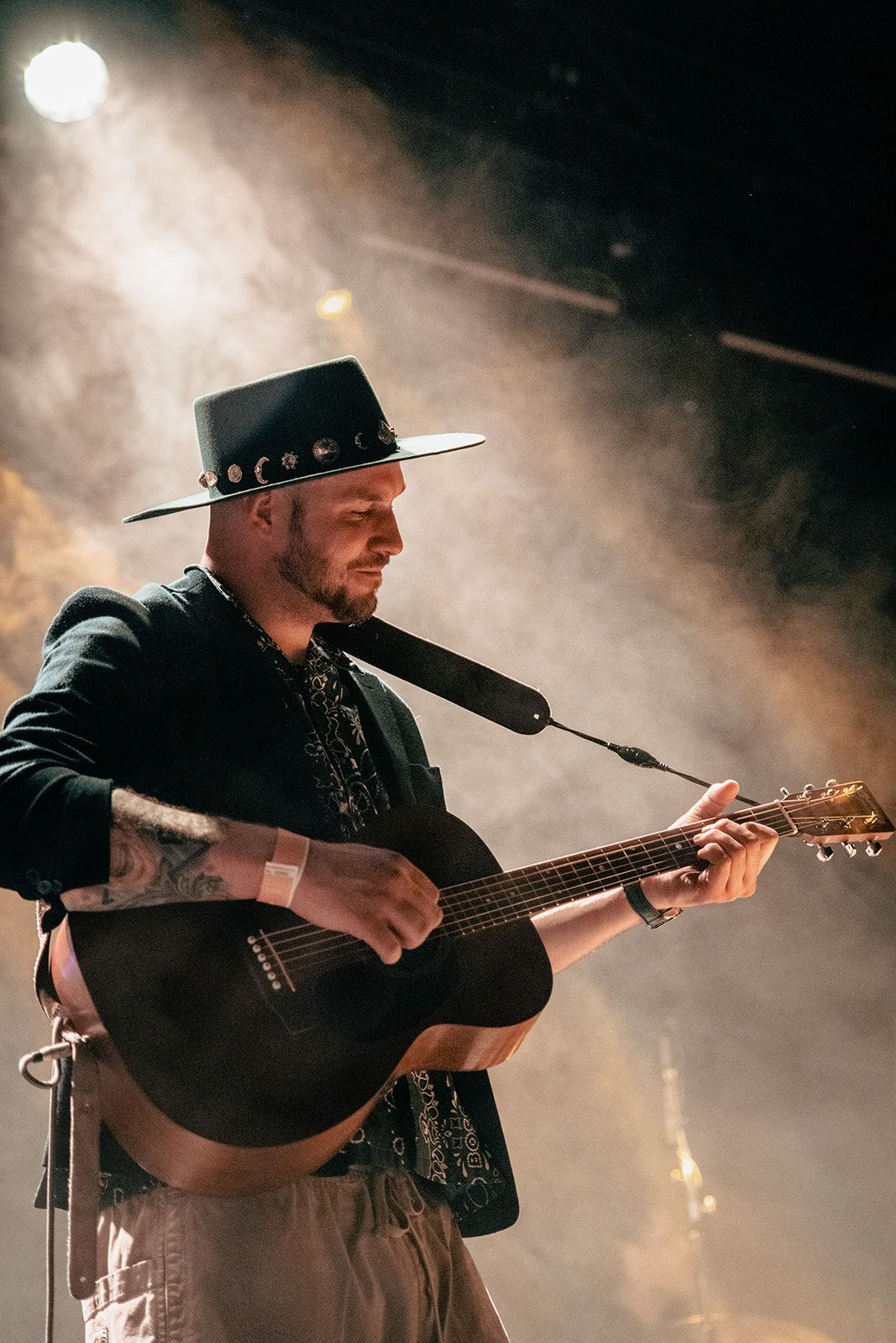 A musician in a black hat performs on acoustic guitar on a smoky stage under dramatic lighting.