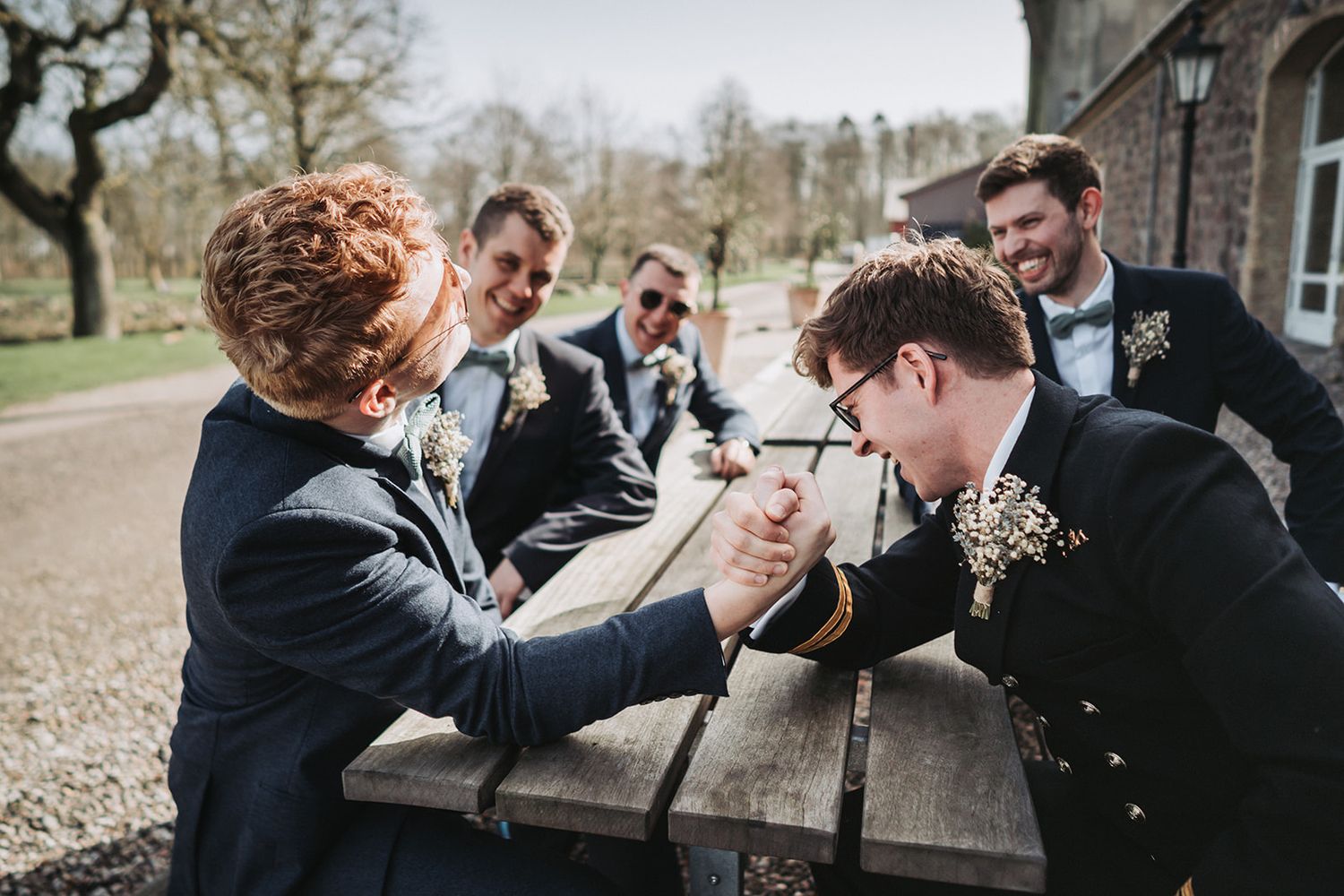 A group wearing dark suits gathers around a wooden picnic table for an arm wrestling match on a cloudy day.