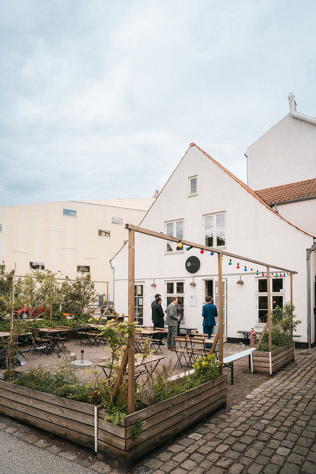 White coastal buildings with raised wooden garden beds and stone paved courtyard create a charming seaside community space.