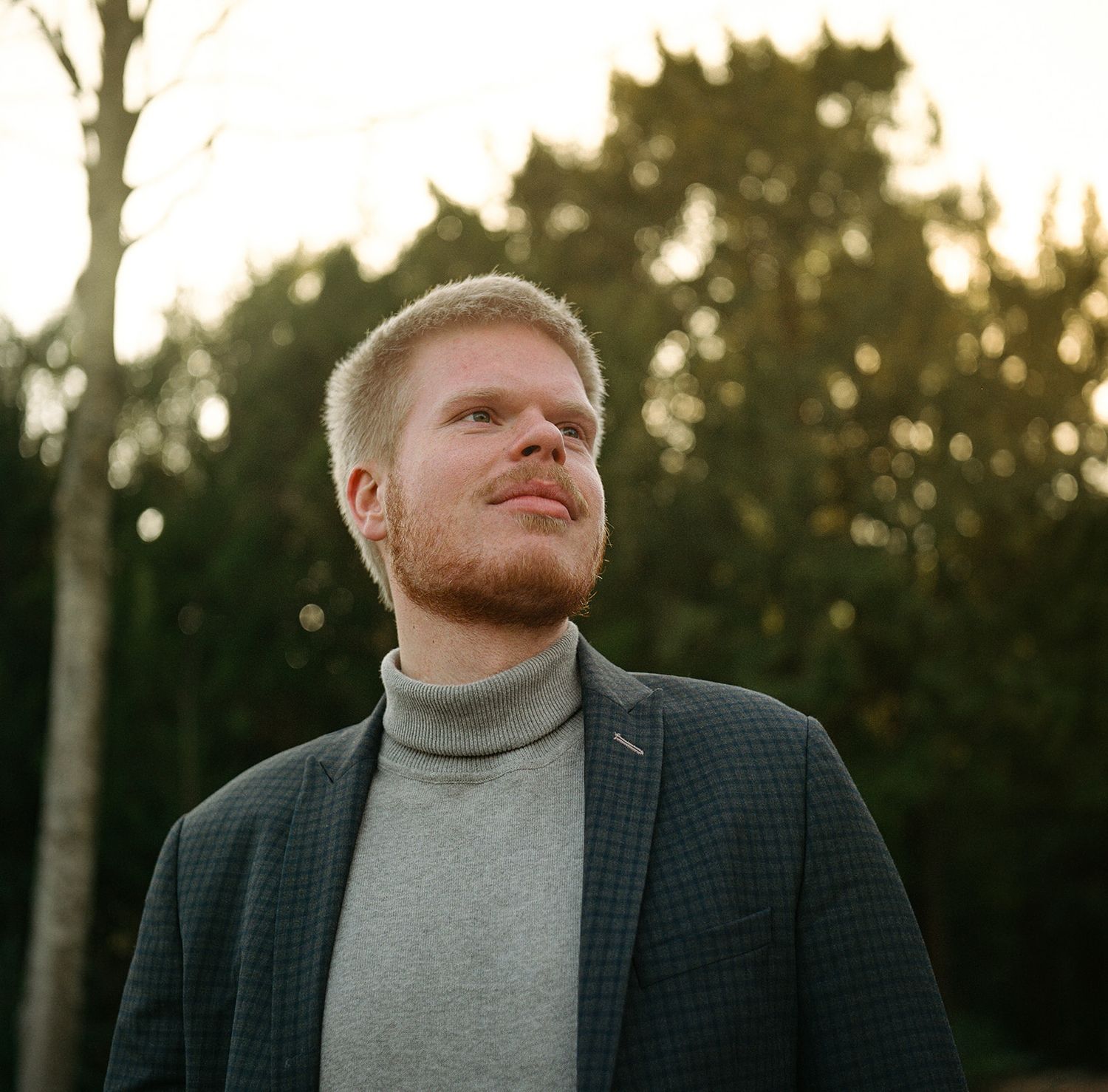 Person in gray turtleneck and dark blazer poses outdoors with soft sunlit background during golden hour.
