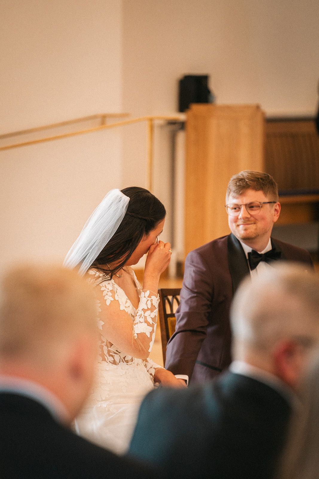 Emotional wedding ceremony moment with bride in lace dress covering face as groom in tuxedo smiles.
