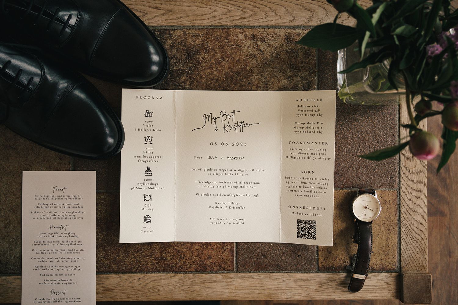 Wedding invitation suite with RSVP cards and envelopes arranged on wooden surface next to dress shoes and greenery.