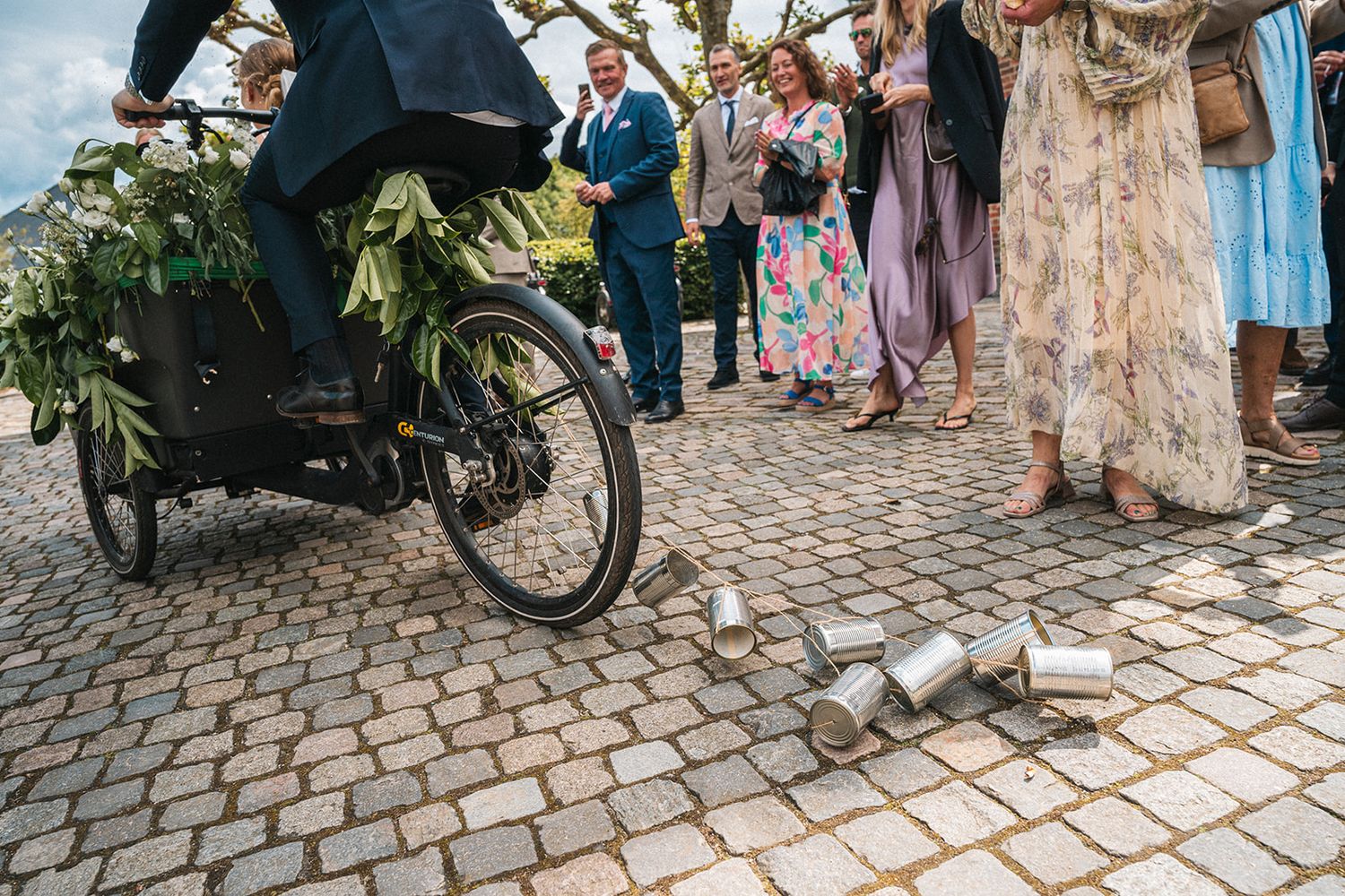 Wedding guests gather on cobblestone street with bicycle and scattered tin cans on the ground during a celebration.