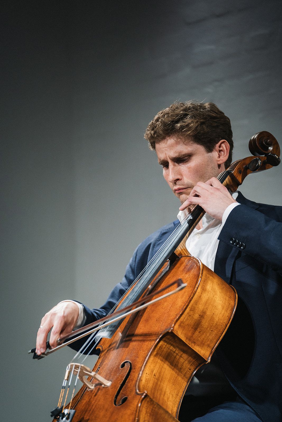 A cellist performs on stage with dramatic dark lighting and plays their wooden string instrument with intense focus and precision.