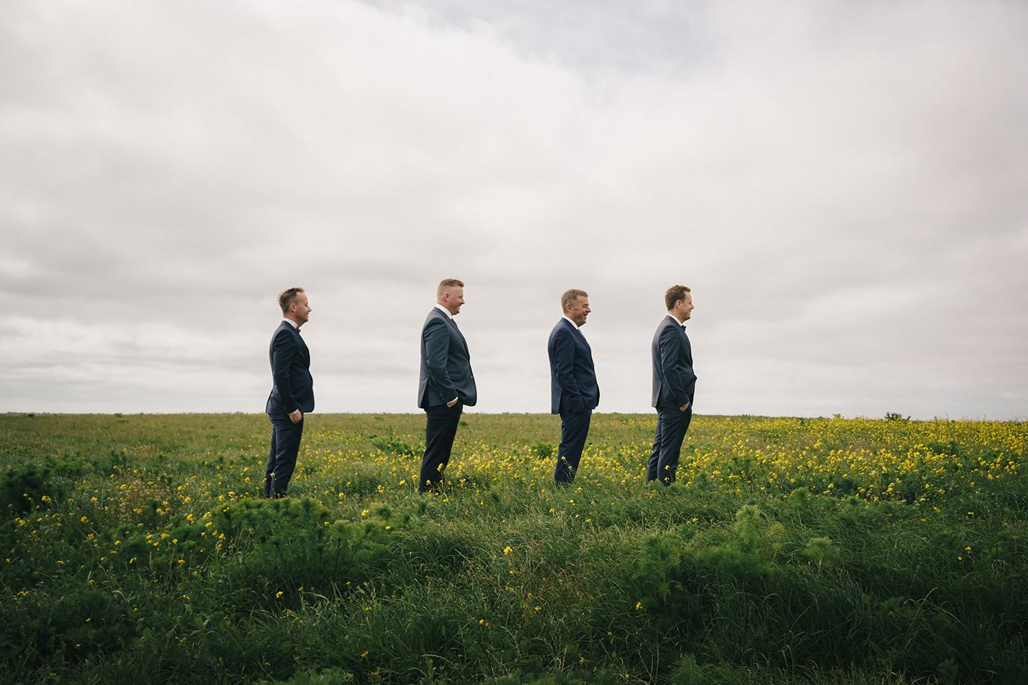 Four groomsmen stand in a line on a grassy hill under an overcast sky.