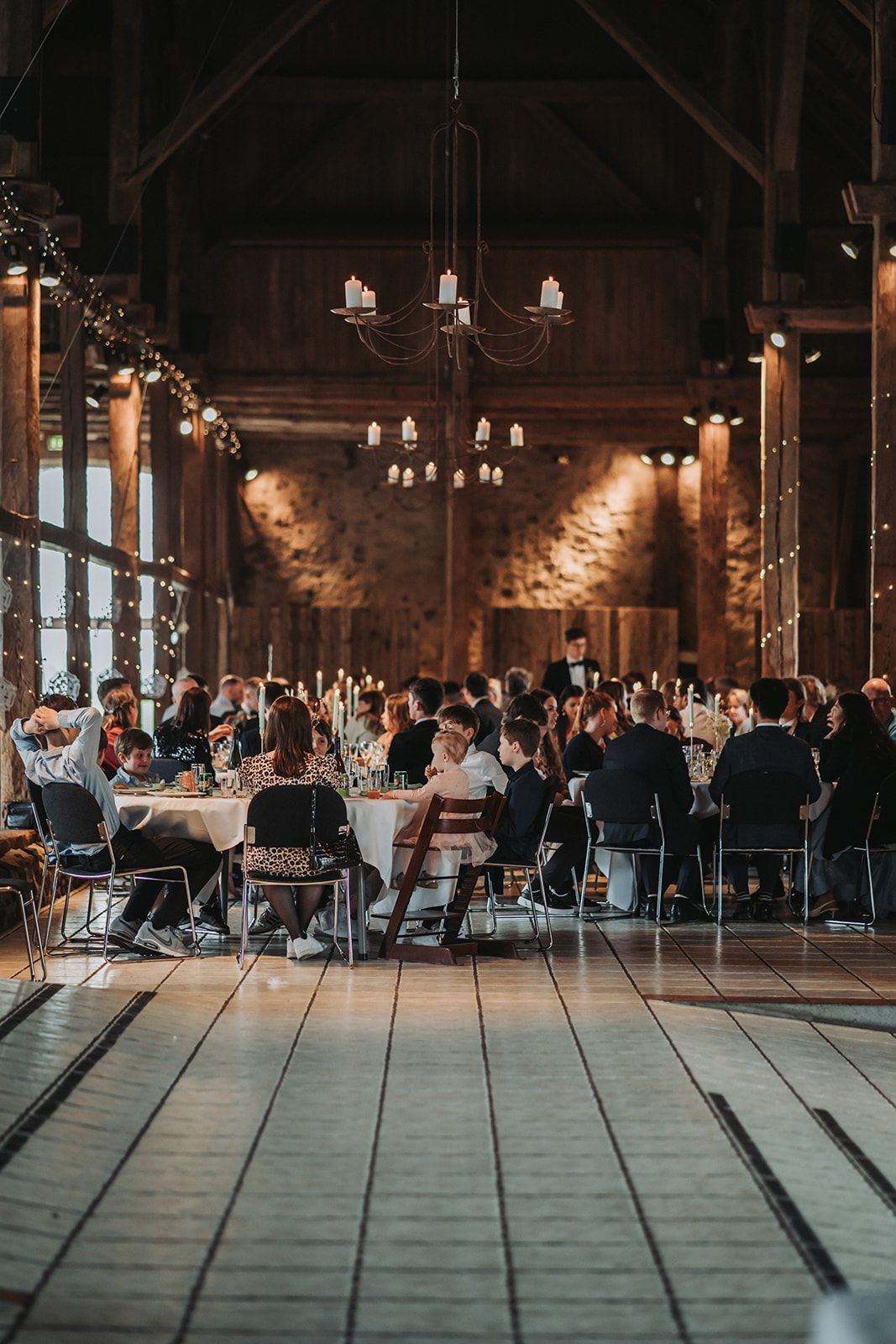 Wedding reception in a rustic industrial venue with hanging lights and guests seated at long tables for dinner.