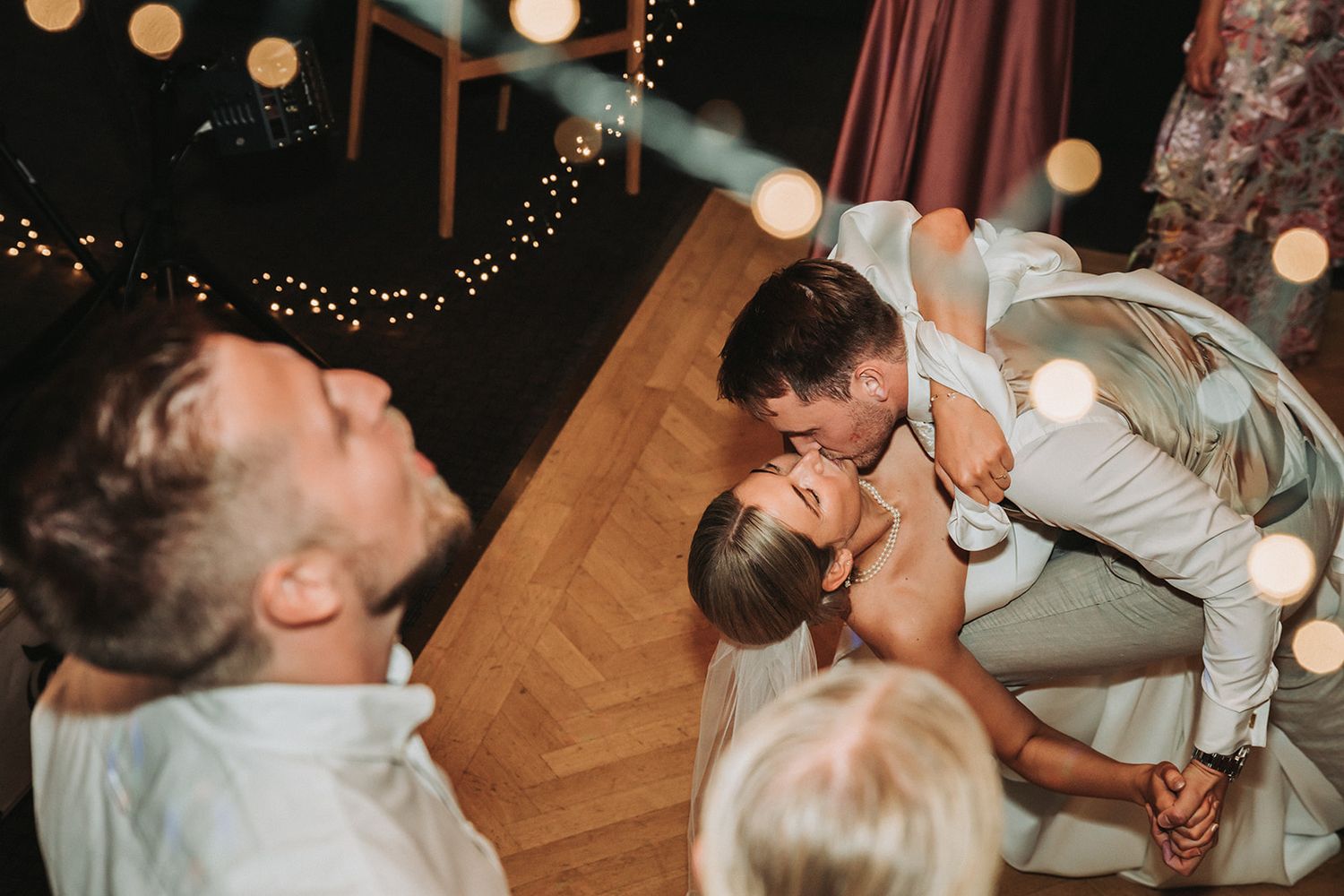 Wedding guests laugh and dance joyfully on a wooden dance floor under warm glowing lights at an evening reception.