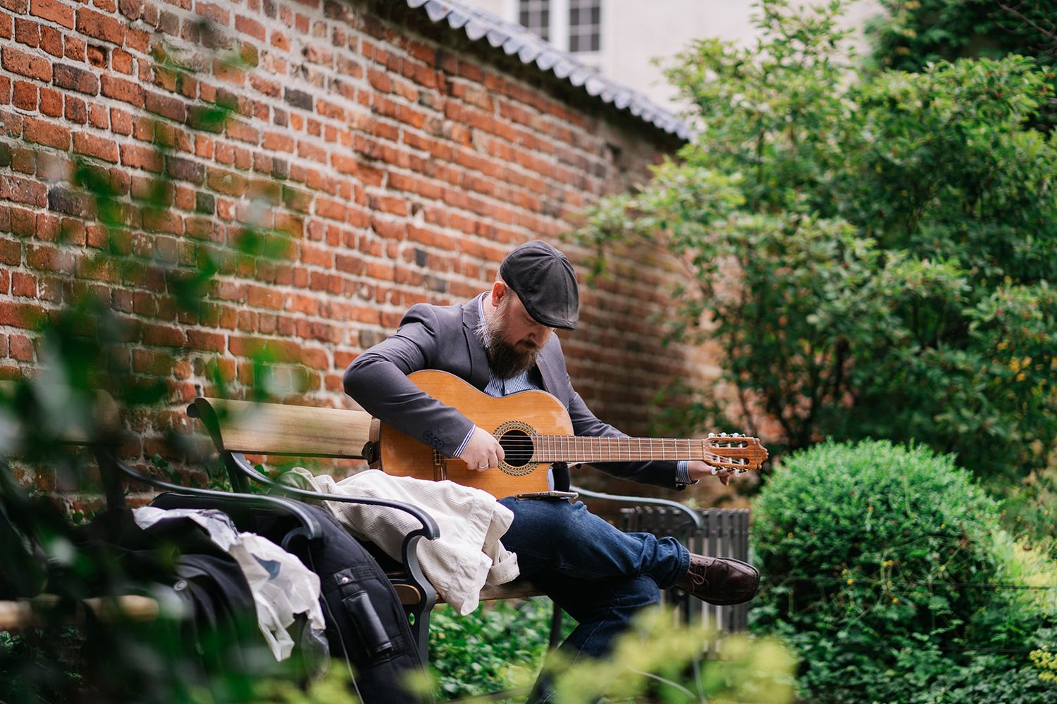 A person in casual attire plays an acoustic guitar while sitting in a peaceful garden with brick wall backdrop.