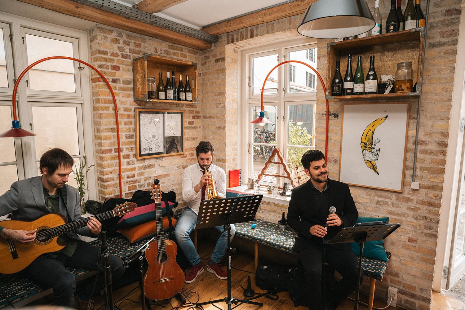 Musicians performing in a rustic brick-walled room with arched windows and acoustic instruments.