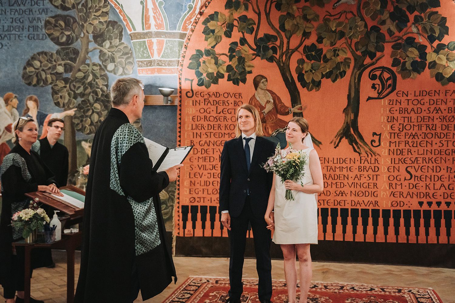 Wedding ceremony in front of vibrant orange folk art mural with decorative trees and text patterns.