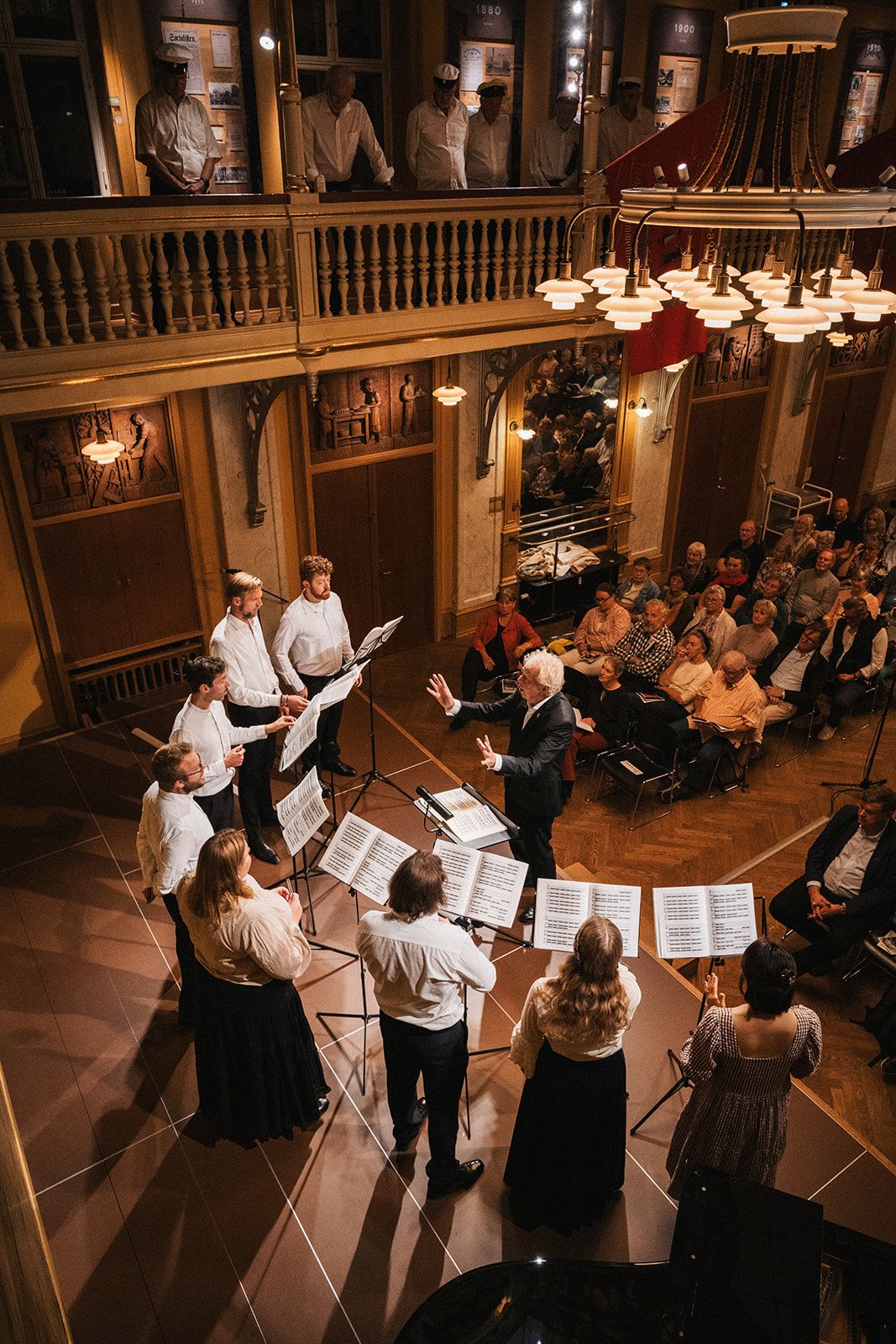 Choir performers in formal attire sing from music stands in a historic wooden concert hall with balcony and audience below.
