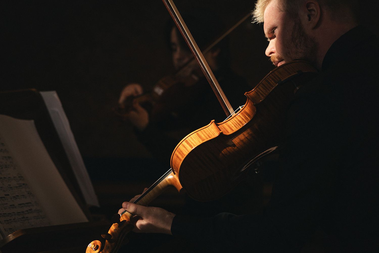 A violin player performs in dramatic lighting while reading sheet music on a stand.