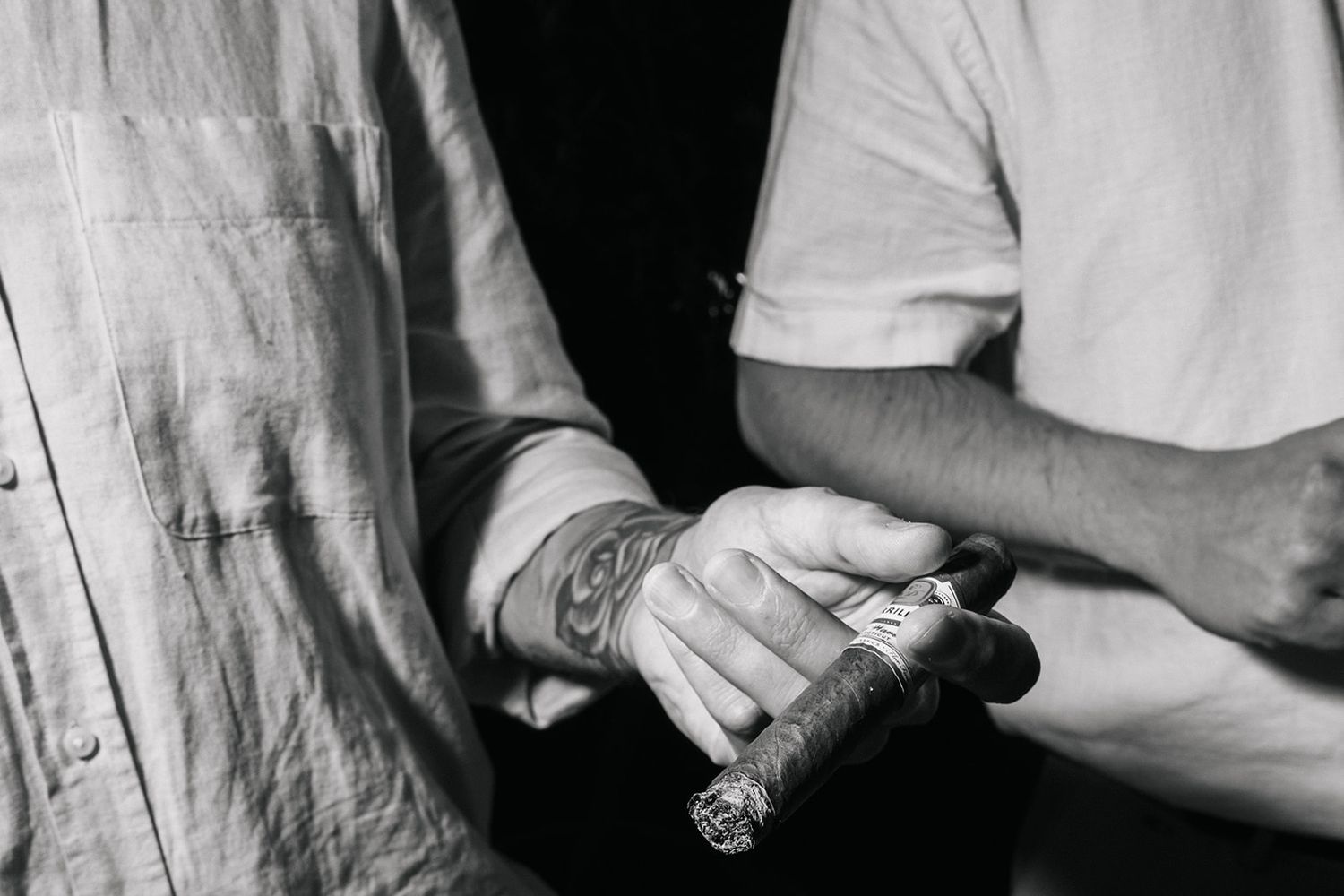 Black and white image of hands passing a cigar between them with tattoo details visible.