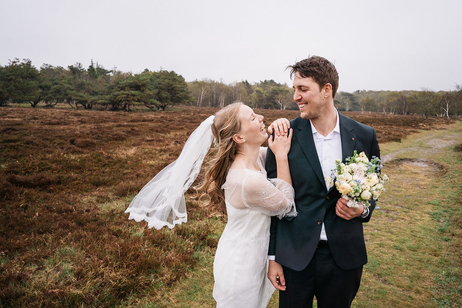 A couple in wedding attire shares joyful moments outdoors with rustic fall scenery in the background.