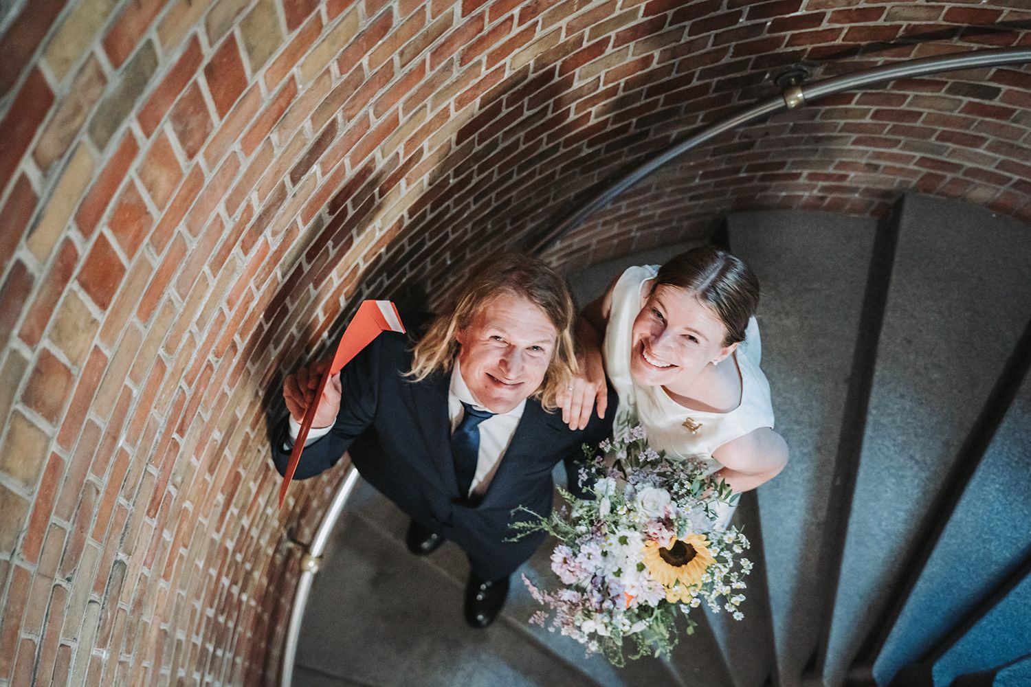 Overhead view of a happy couple on their wedding day standing on spiral staircase with brick walls and floral bouquet.
