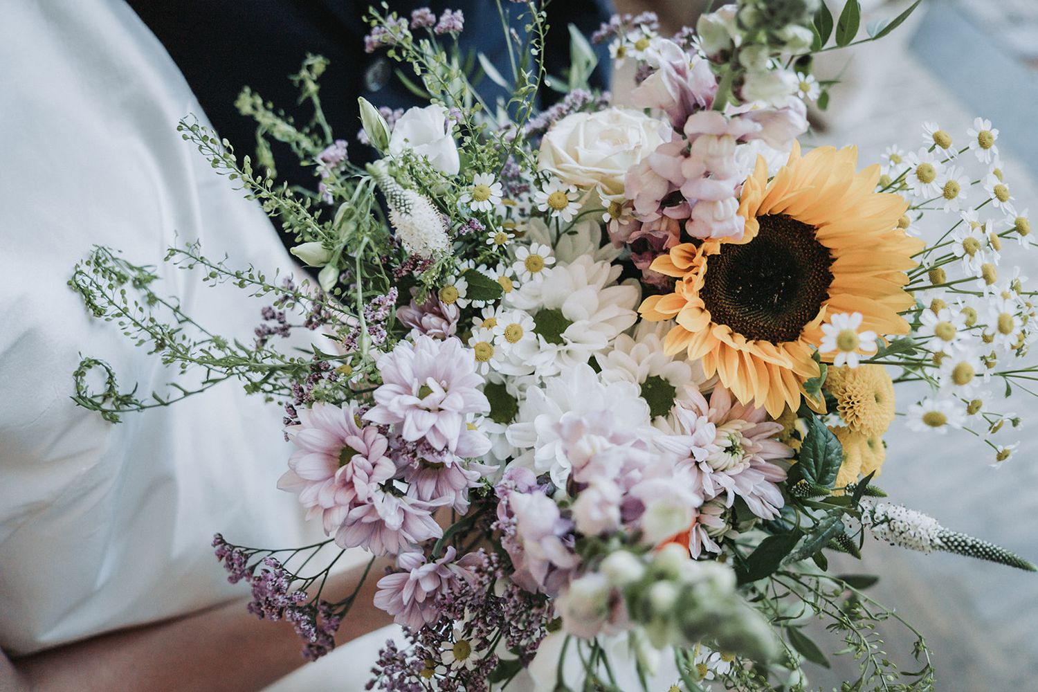A rustic floral bouquet featuring a bright sunflower, pink blooms, and delicate baby's breath against white fabric.