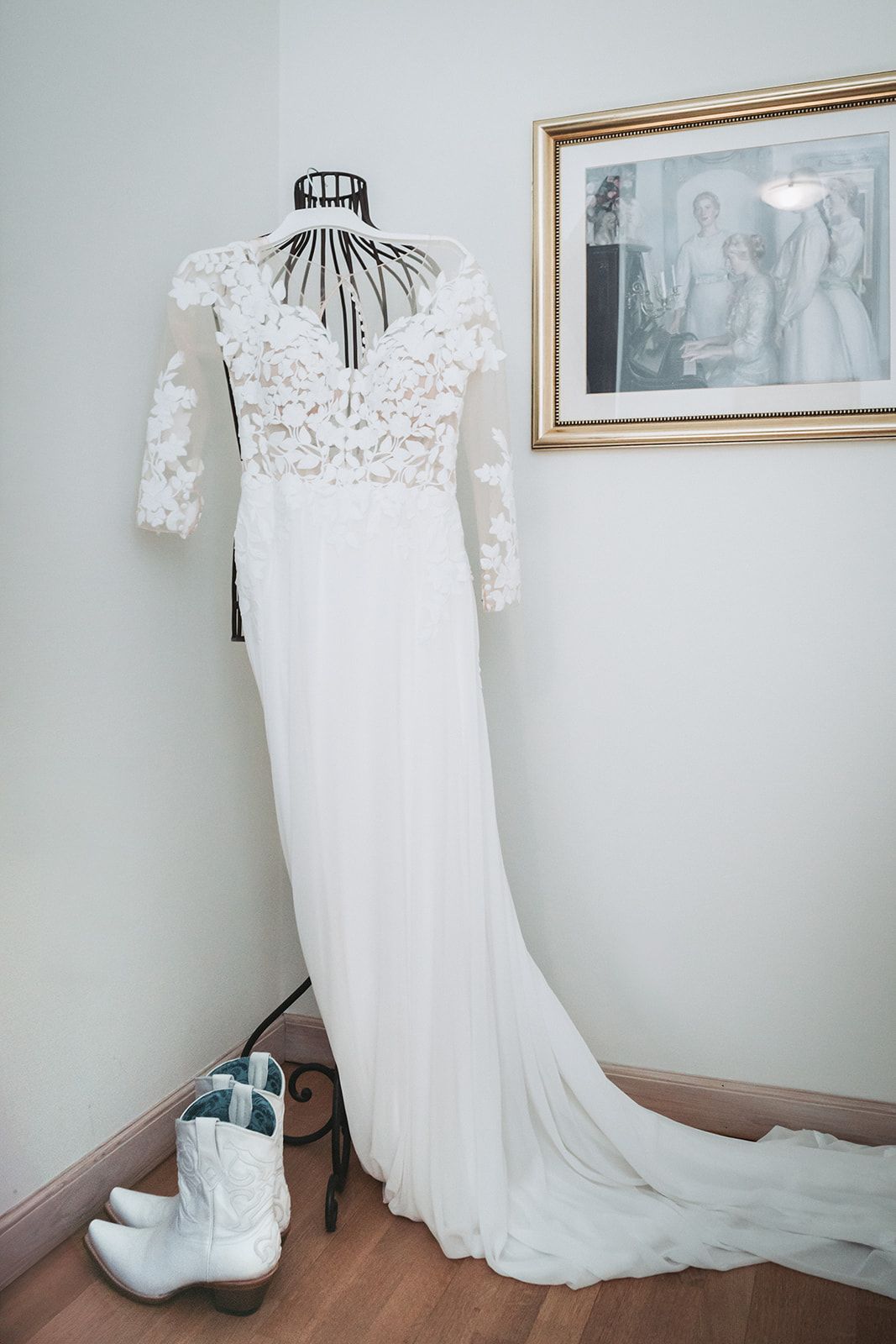 A white lace wedding dress with long sleeves and train hangs on a dress form with white cowboy boots below it.
