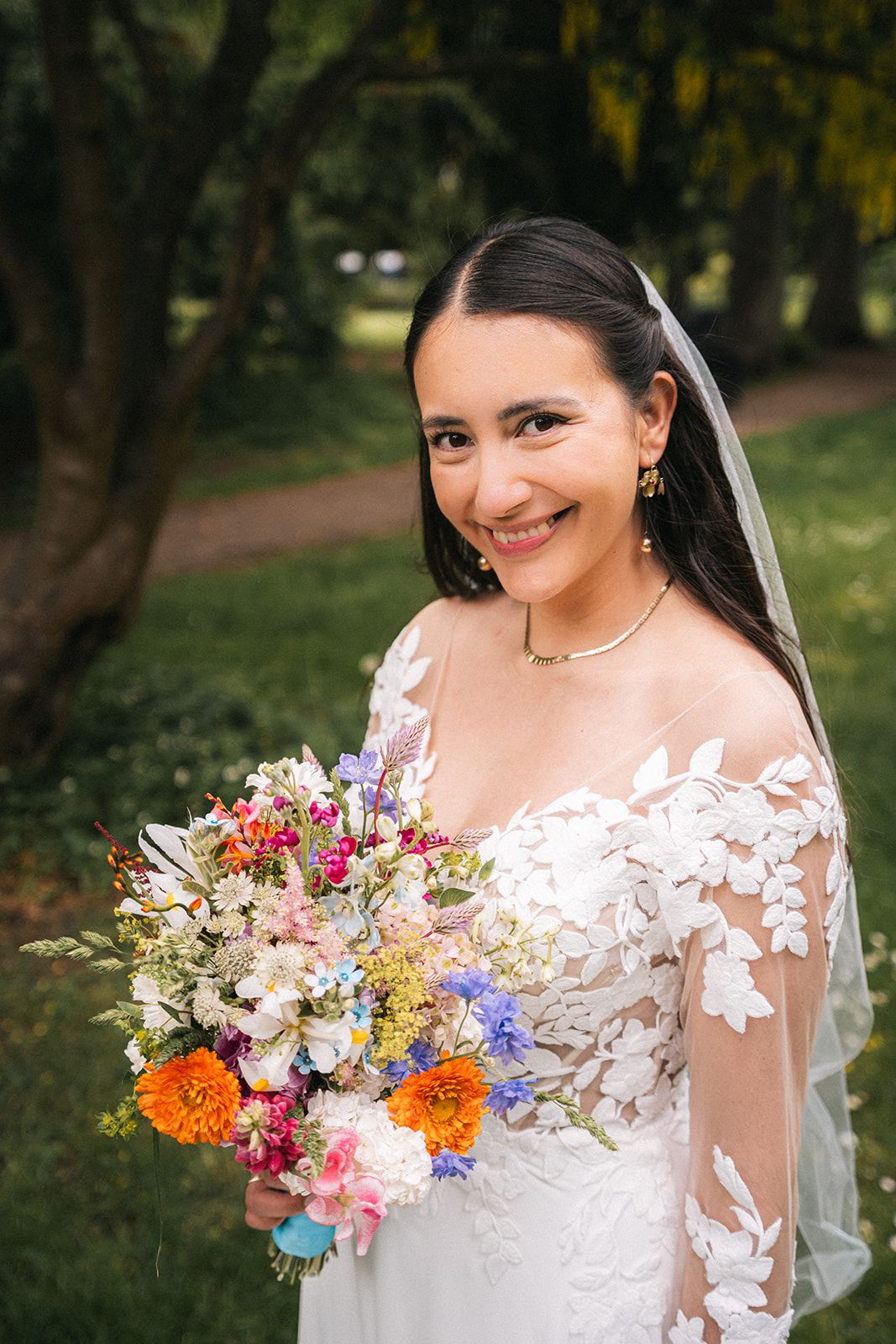 A joyful bride in a white lace dress holds a vibrant wildflower bouquet featuring orange, pink and blue blooms.