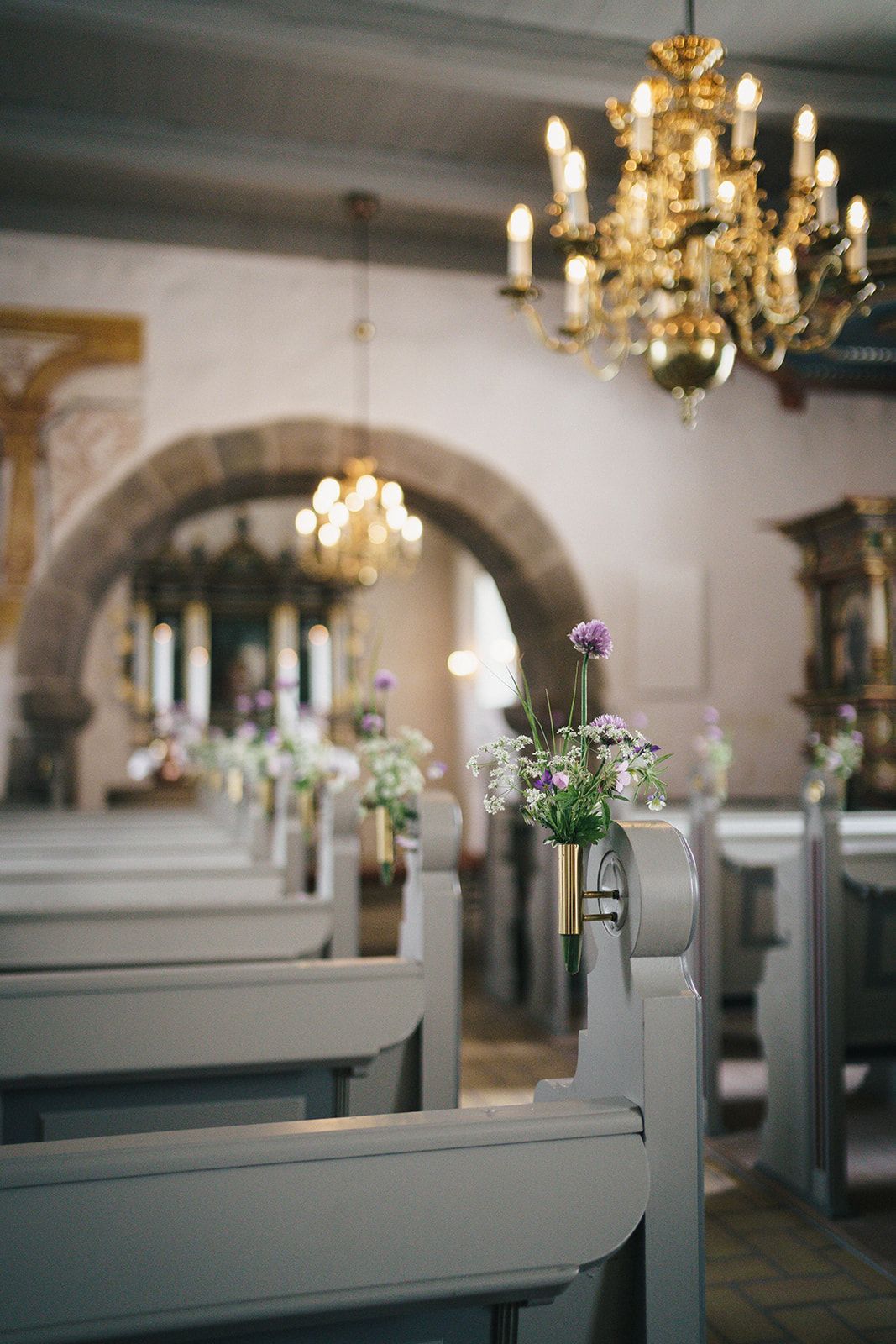 Interior of a church with gray wooden pews decorated with delicate flower arrangements and illuminated by brass chandeliers.
