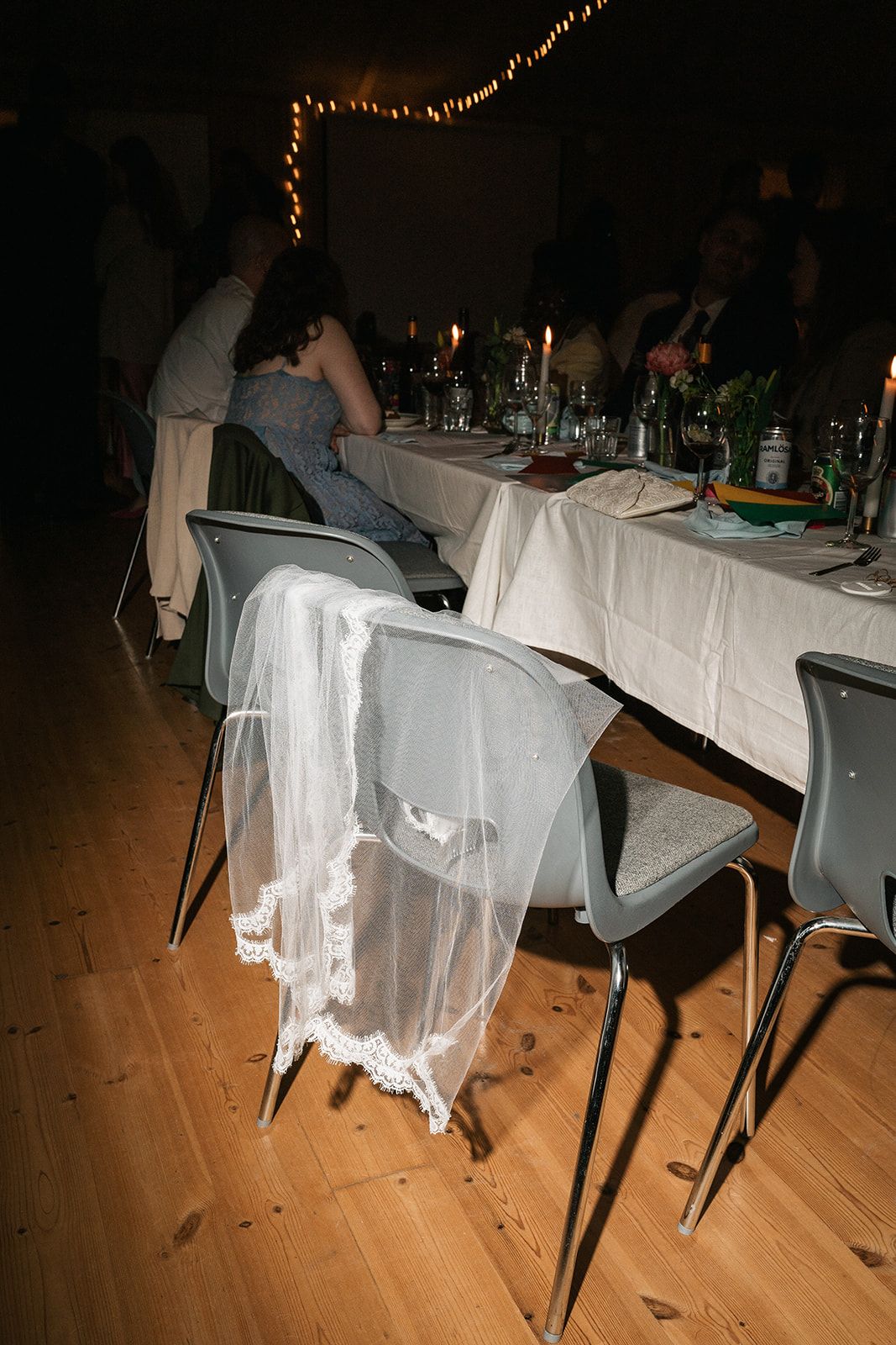 A dining table setup at a party with chairs draped in white fabric and string lights overhead on a wooden floor.