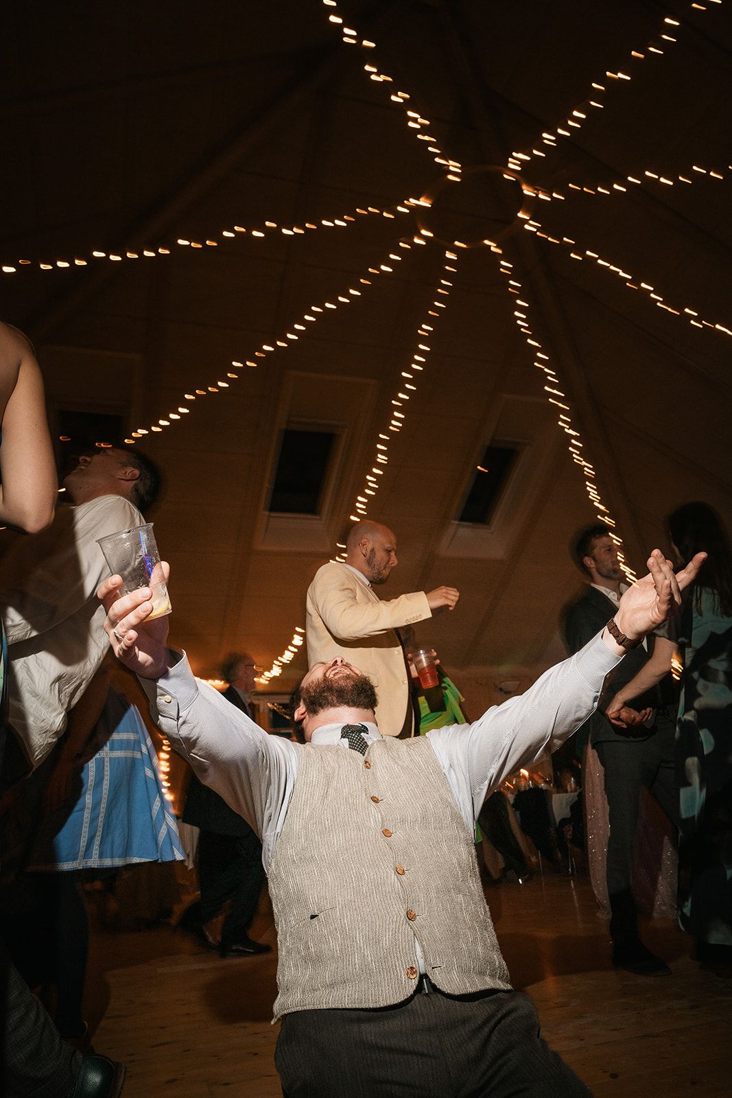 Wedding guest dances enthusiastically with arms raised under decorative string lights at indoor reception venue.
