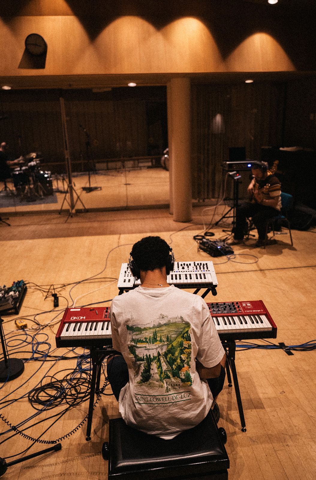 Musician seen from behind playing electronic keyboards during a recording session in a warm-lit studio space.