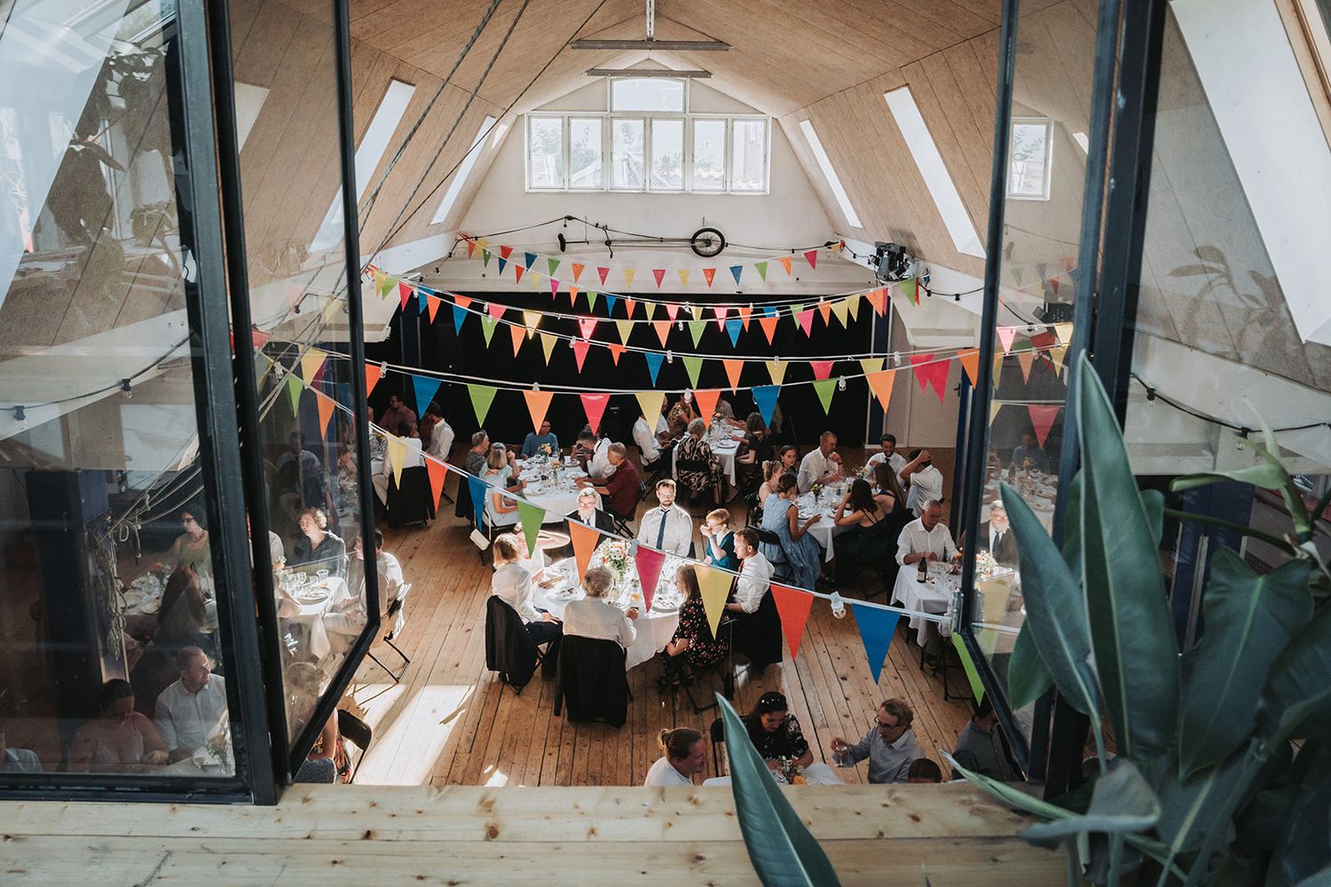 Wedding reception venue decorated with colorful bunting flags and guests seated at round tables in a rustic attic space.