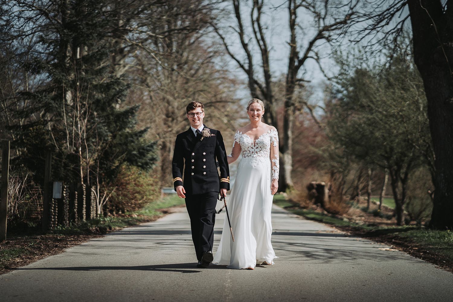 A couple walking together on a tree-lined path, with the person on the left in military dress and right in white gown.