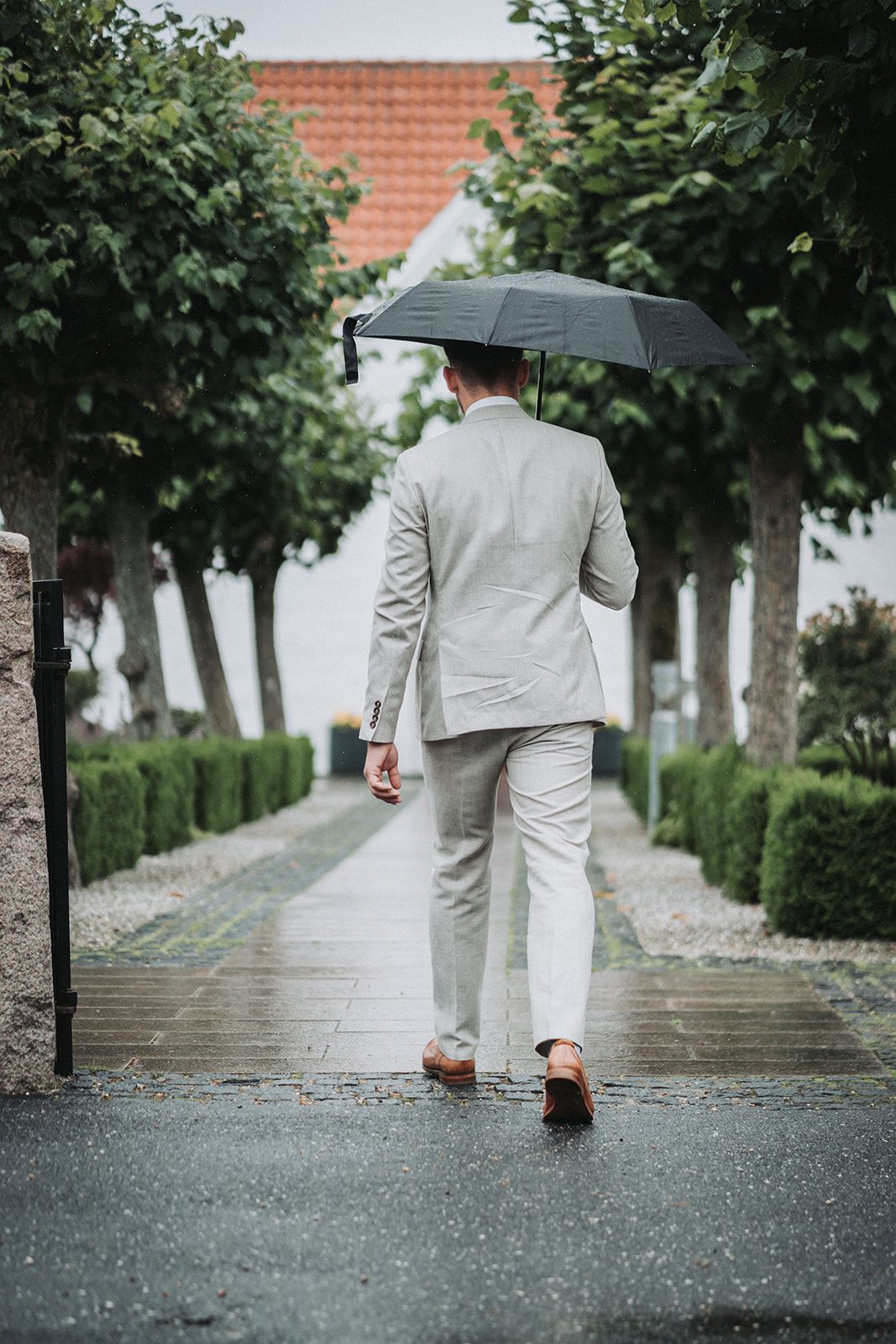 A gentleman in a light gray suit holds a black umbrella while walking on a rainy path lined with trees.