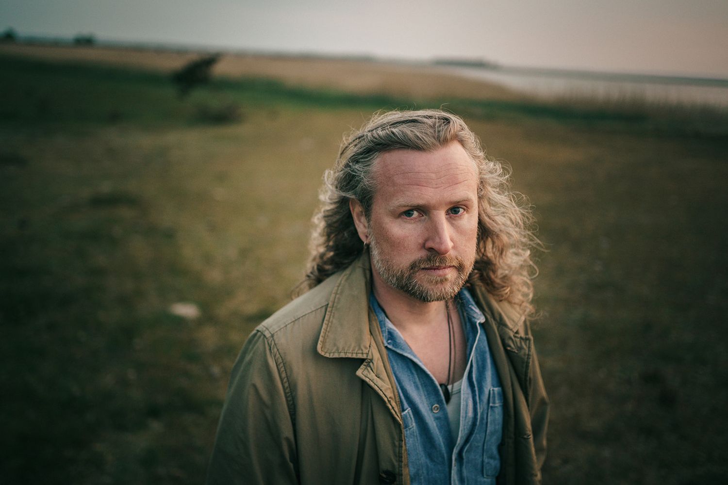A person with long blonde hair and a brown jacket stands in a field during golden hour.