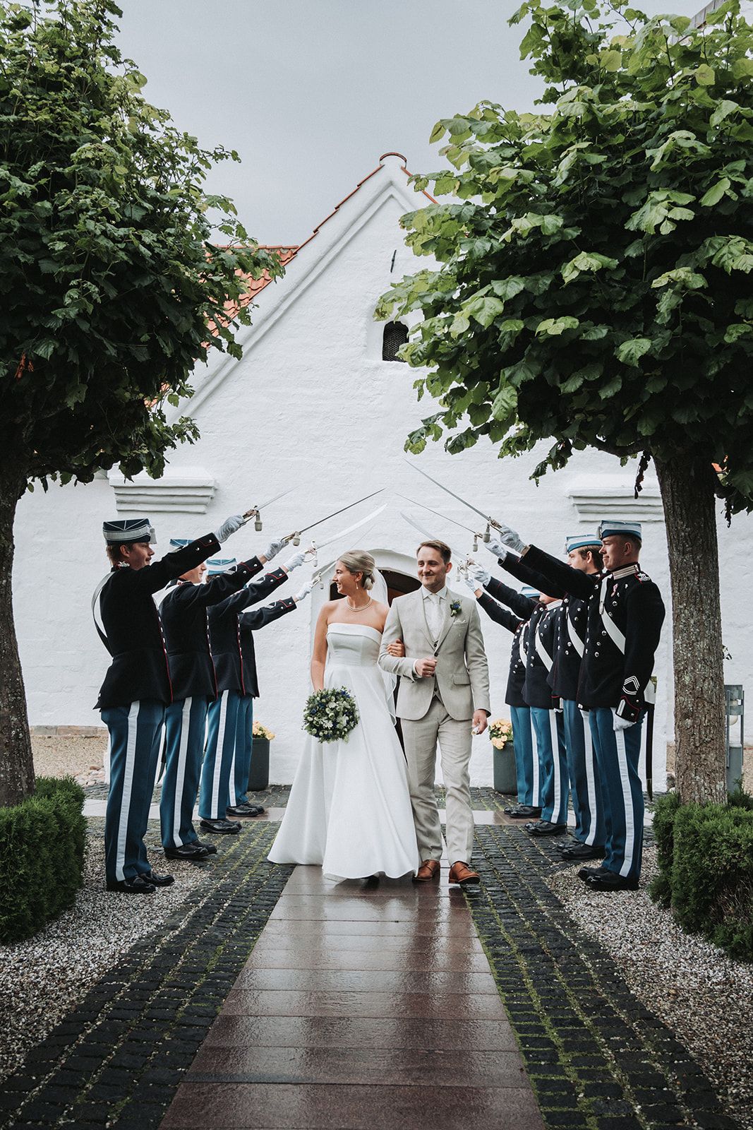 A wedding party walks under trees while groomsmen in blue uniforms create an arch against a white building backdrop.