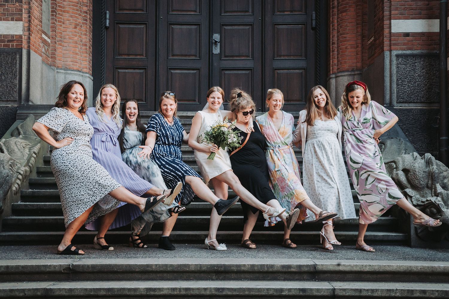 Bridesmaids in pastel dresses striking playful poses on stone steps.
