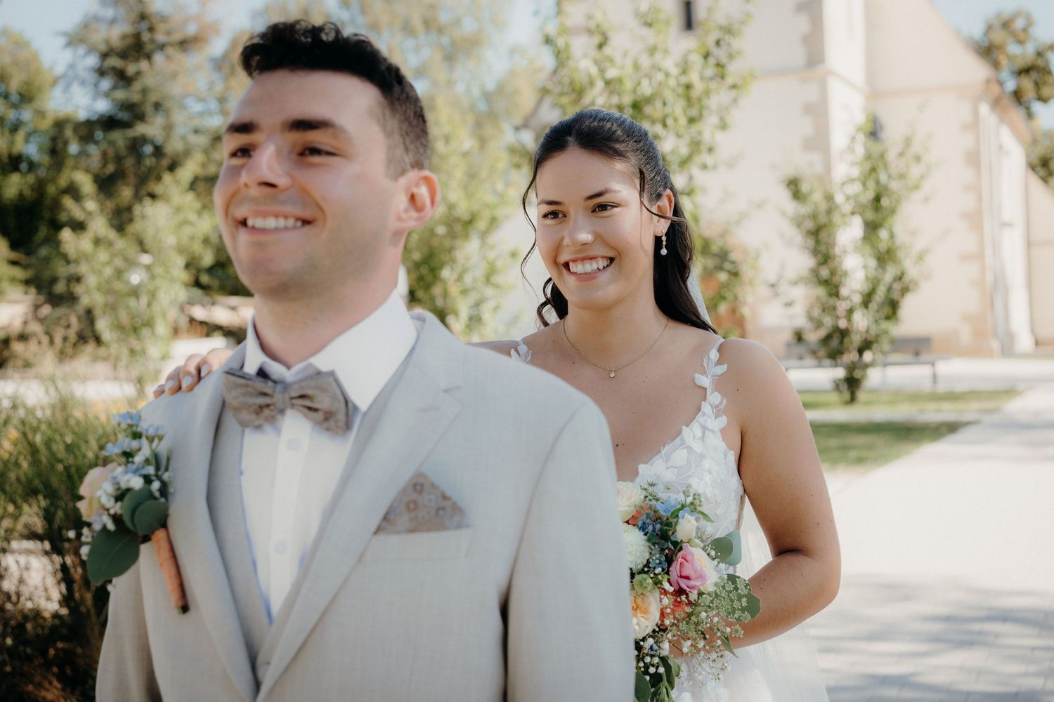 A smiling bride in a white dress holds a floral bouquet beside her groom in a beige suit with a bow tie outdoors.