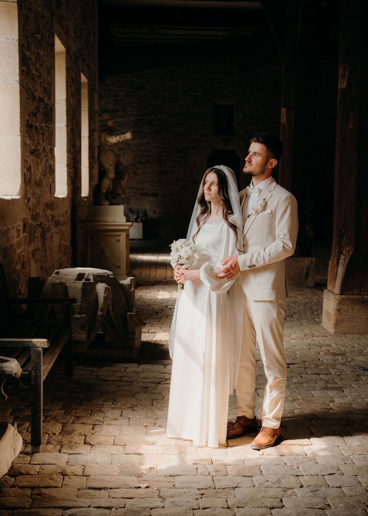 Bride and groom stand in soft light inside a rustic stone venue, bride holding white bouquet in elegant wedding attire.