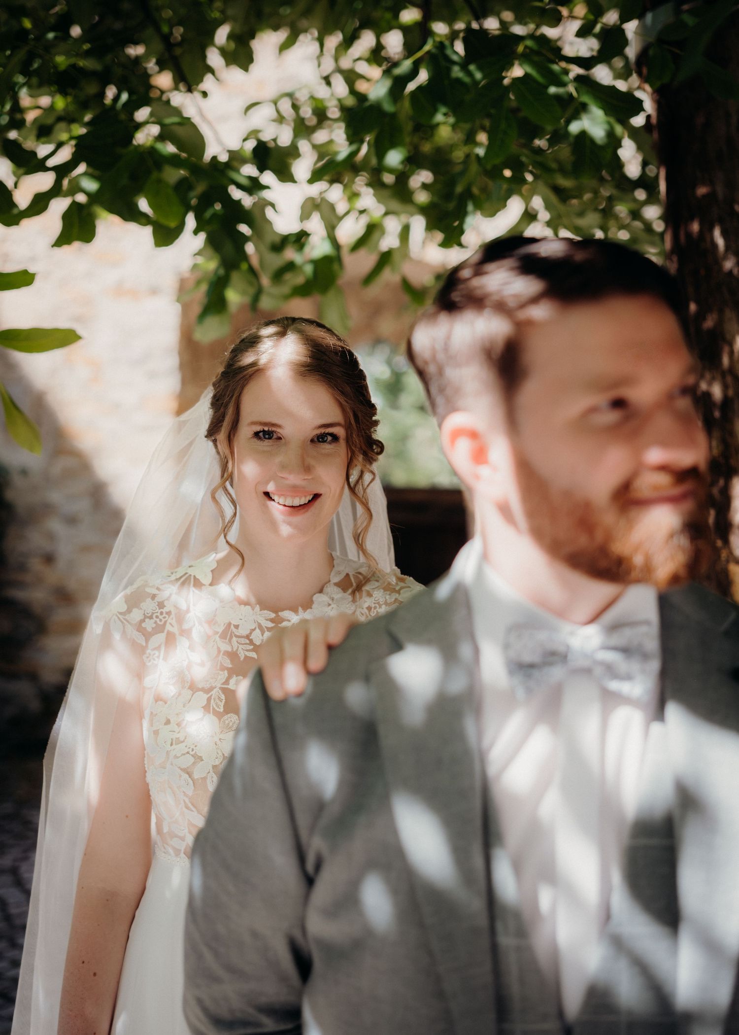 A smiling bride in lace gown and veil stands behind a bearded groom in gray suit with bow tie under leafy trees.