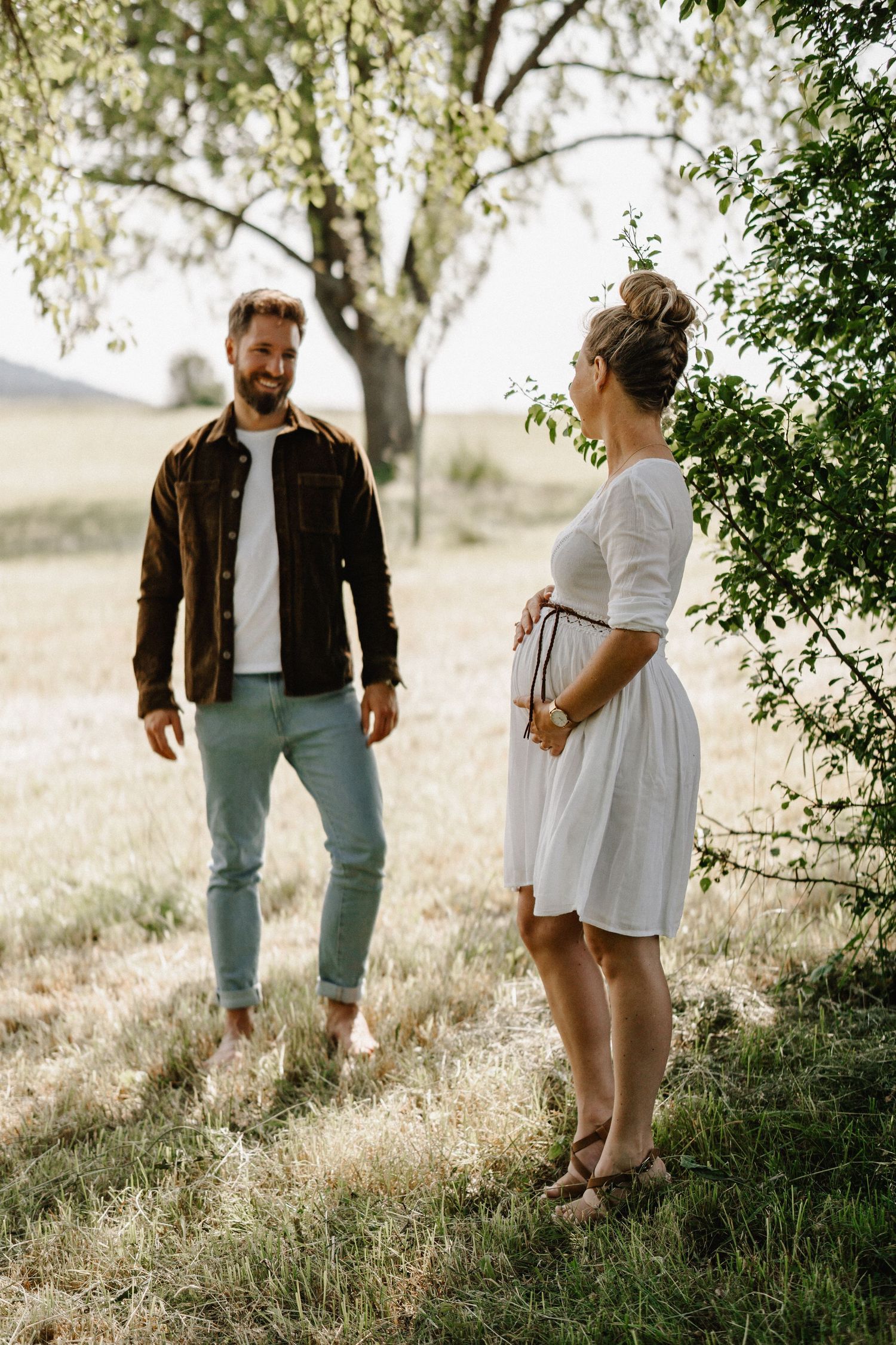 A couple stands together in a sunlit meadow under a tree during a romantic outdoor photoshoot.