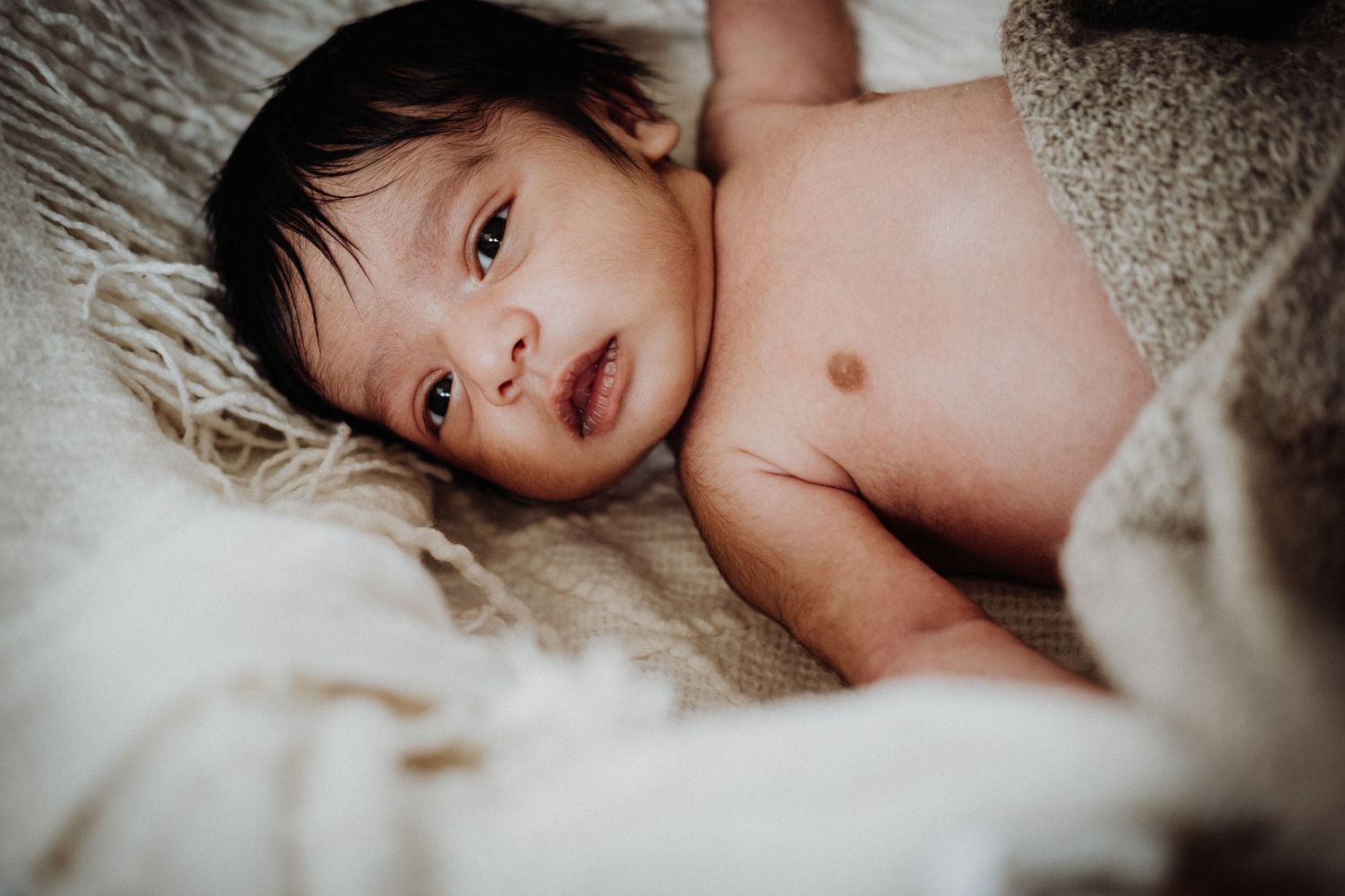 A baby lies peacefully on a soft beige blanket while looking at the camera.